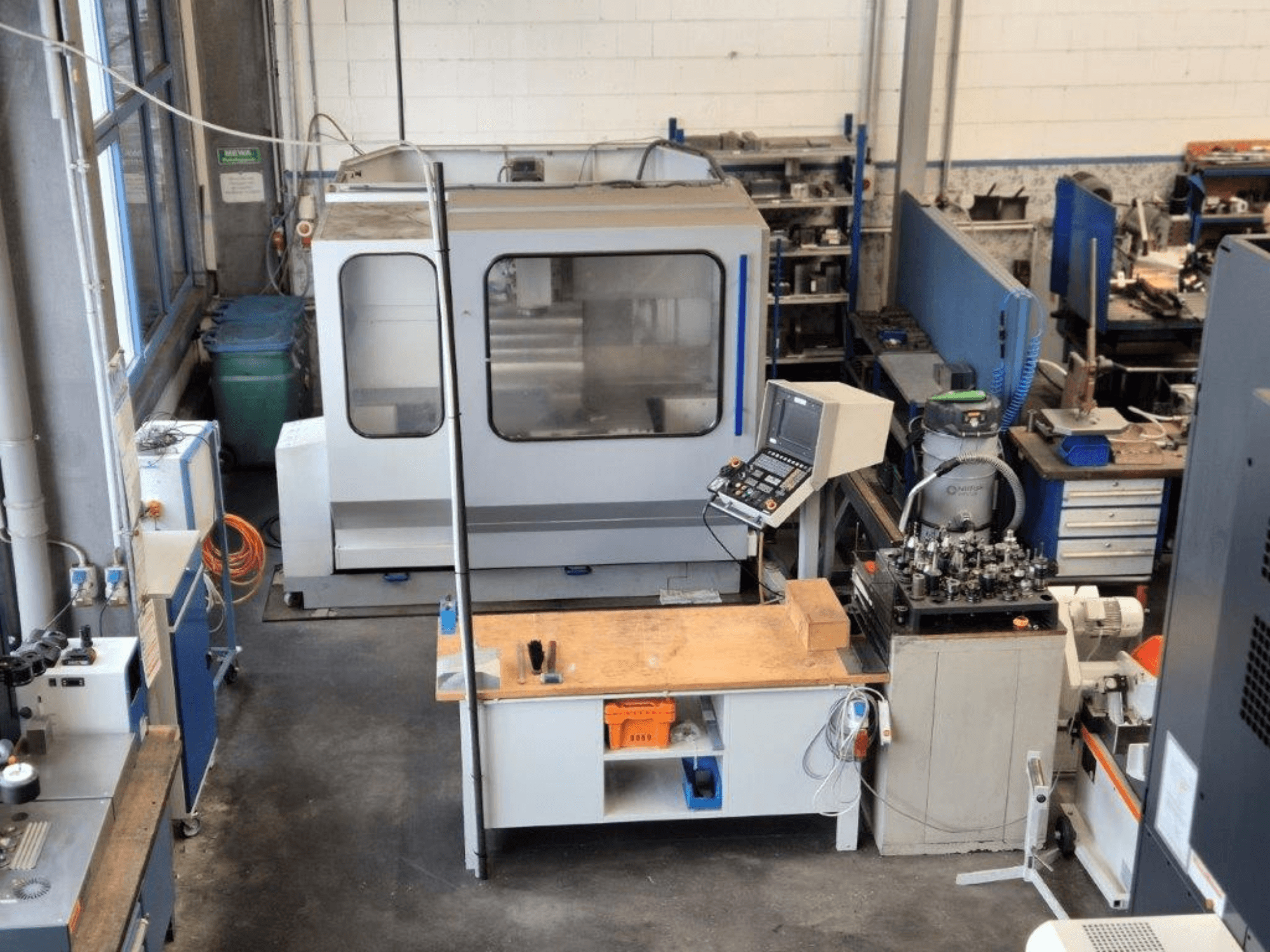 CNC frezavimo staklės dirbtuvėse, vaizdas iš priekio, fone matomas valdymo pultas, darbo paviršius ir įrankiai.