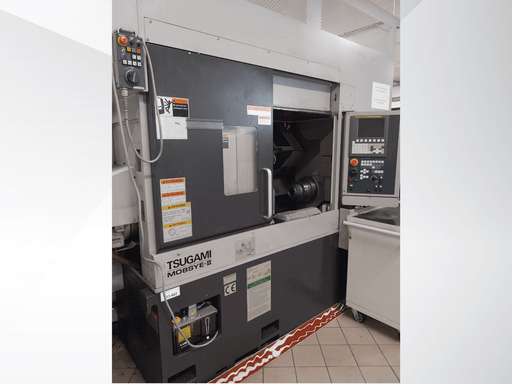 Tsugami M08SY-II CNC tekinimo staklės, priekinis vaizdas, kuriame matyti valdymo skydelis, suklys ir krovimo zona. Yra saugos užrašai ir prekės ženklo etiketė.