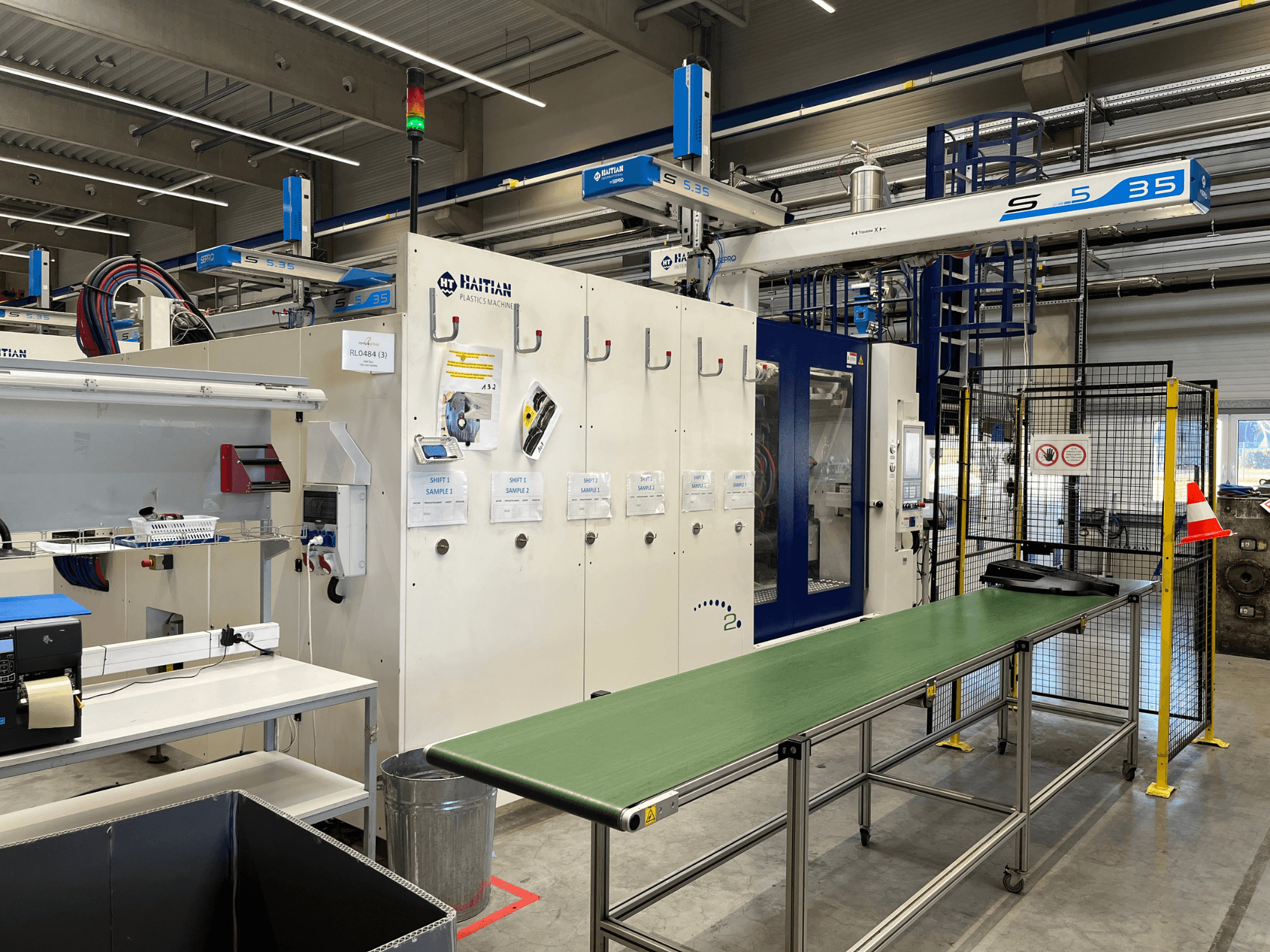 Haičio S5-35 plastiko mašina dirbtuvėse; iš priekio matomas žalias konvejerio stalas, apsauginiai barjerai ir valdymo pultas.