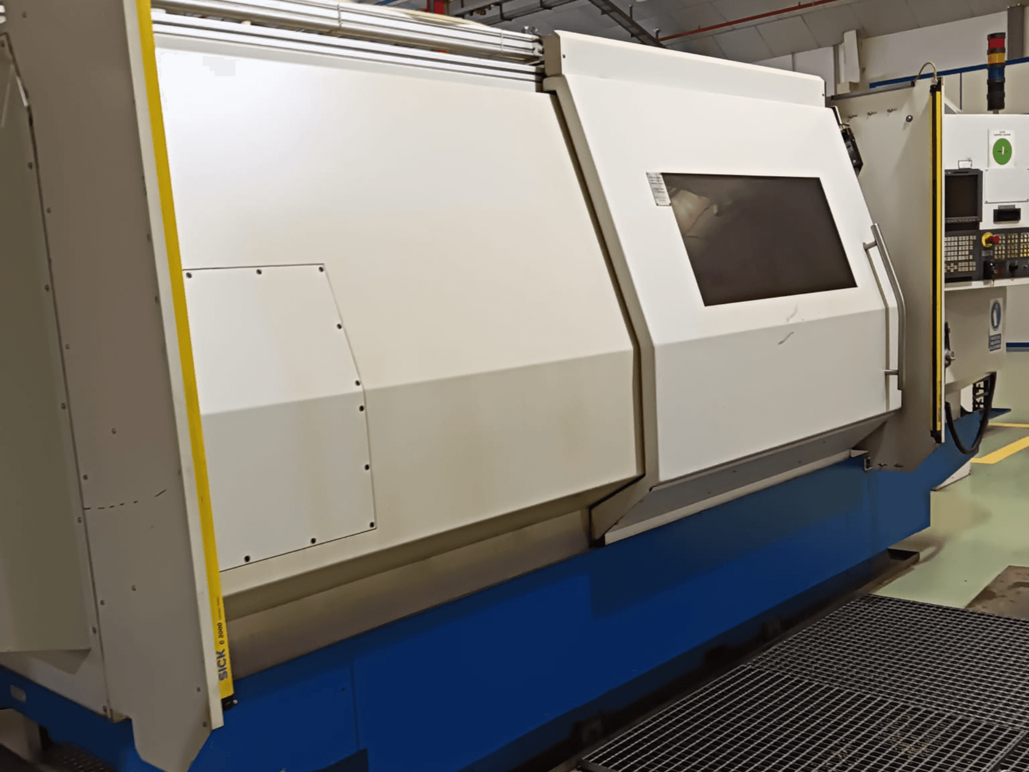 Pramoninės tekinimo staklės, vaizdas iš šono, su dideliu baltos ir mėlynos spalvos korpusu, skaitmeniniu valdymo skydeliu ir saugos funkcijomis.