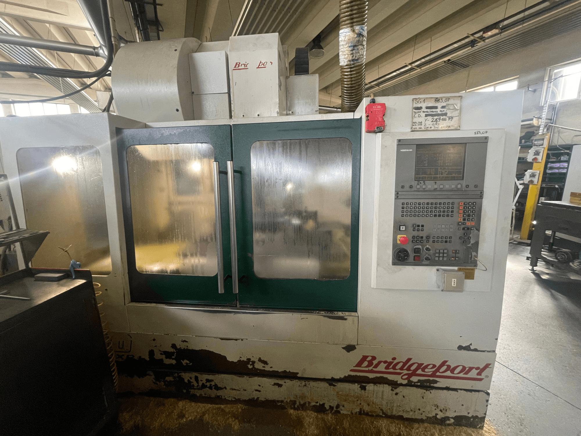 Vertikalus apdirbimo centras „Bridgeport VMC 1000/30“, vaizdas iš priekio su valdymo pultu, matyti nusidėvėjęs išorinis paviršius ir žalios durys.