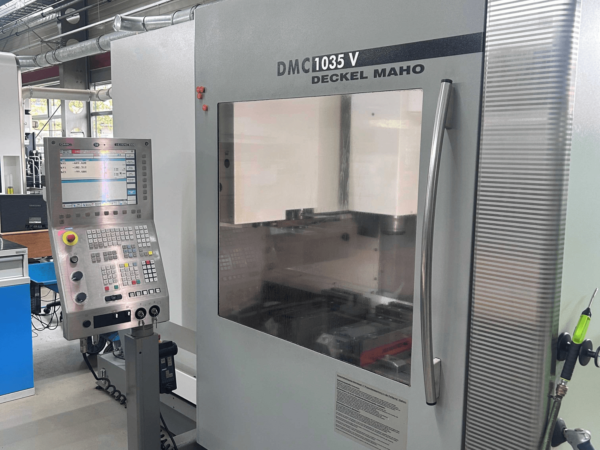 Vertikalaus apdirbimo centro „DECKEL MAHO DMC 1035 V“ vaizdas iš priekio, kuriame matomas valdymo pultas ir skaidrus langas į darbo zoną.