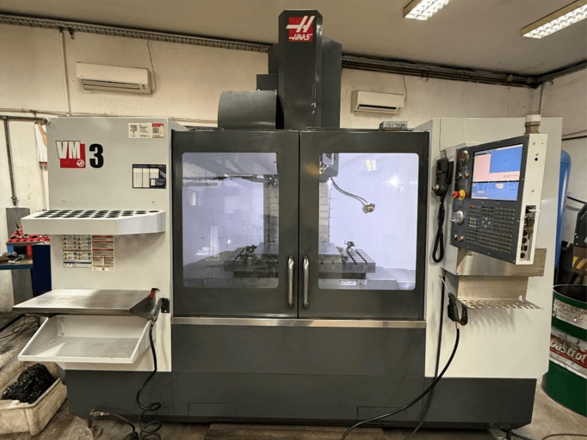 „Haas VM-3“ vertikalaus apdirbimo centro vaizdas iš priekio, kuriame matomas didelis valdymo skydas ir apdirbimo zona su įrankiais.