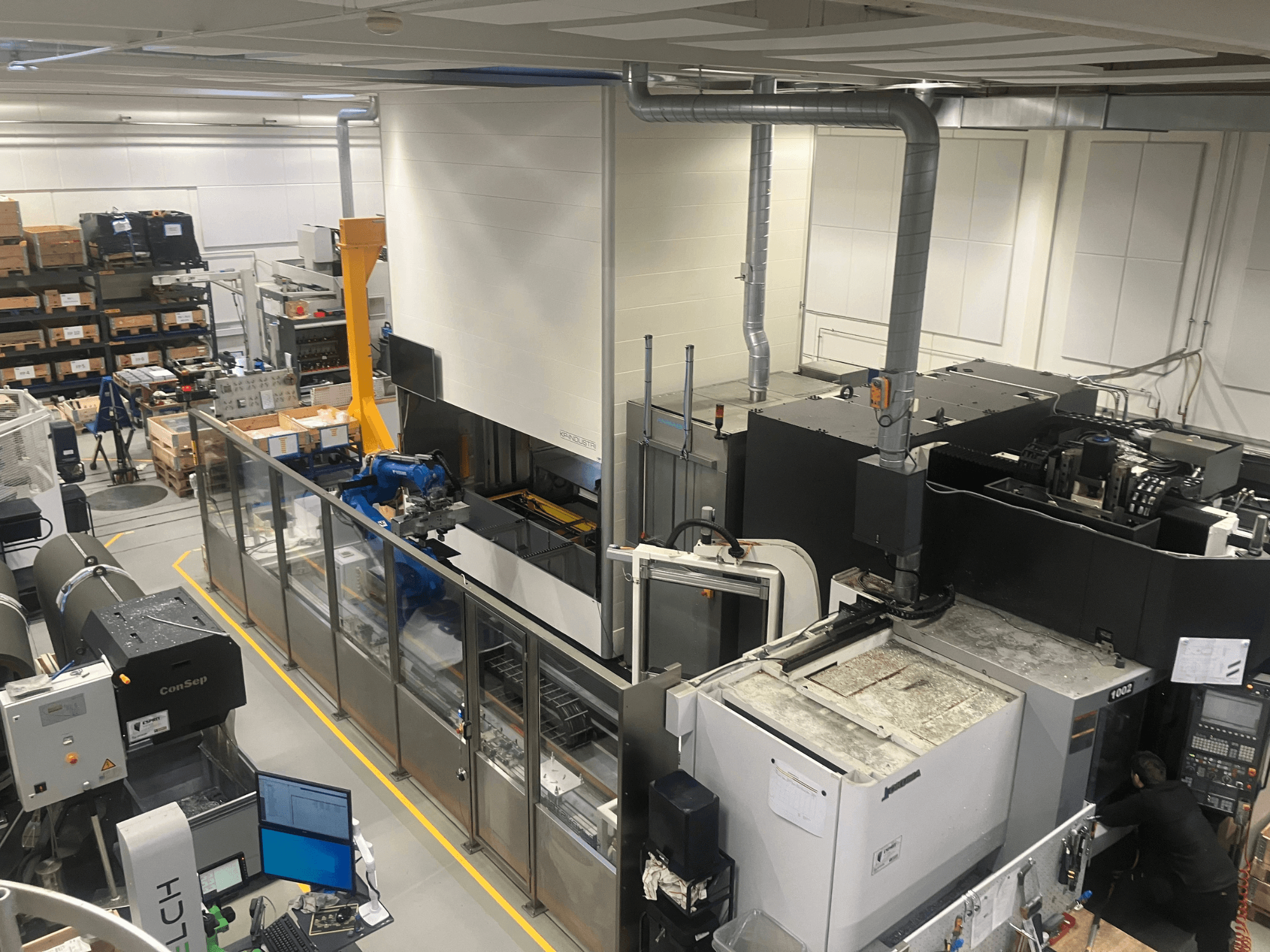 SSI SCHAFER "Logimat" automatizavimo sistema sandėlyje, vaizdas iš priekio; roboto ranka, sandėliavimo stelažai ir mašinos.