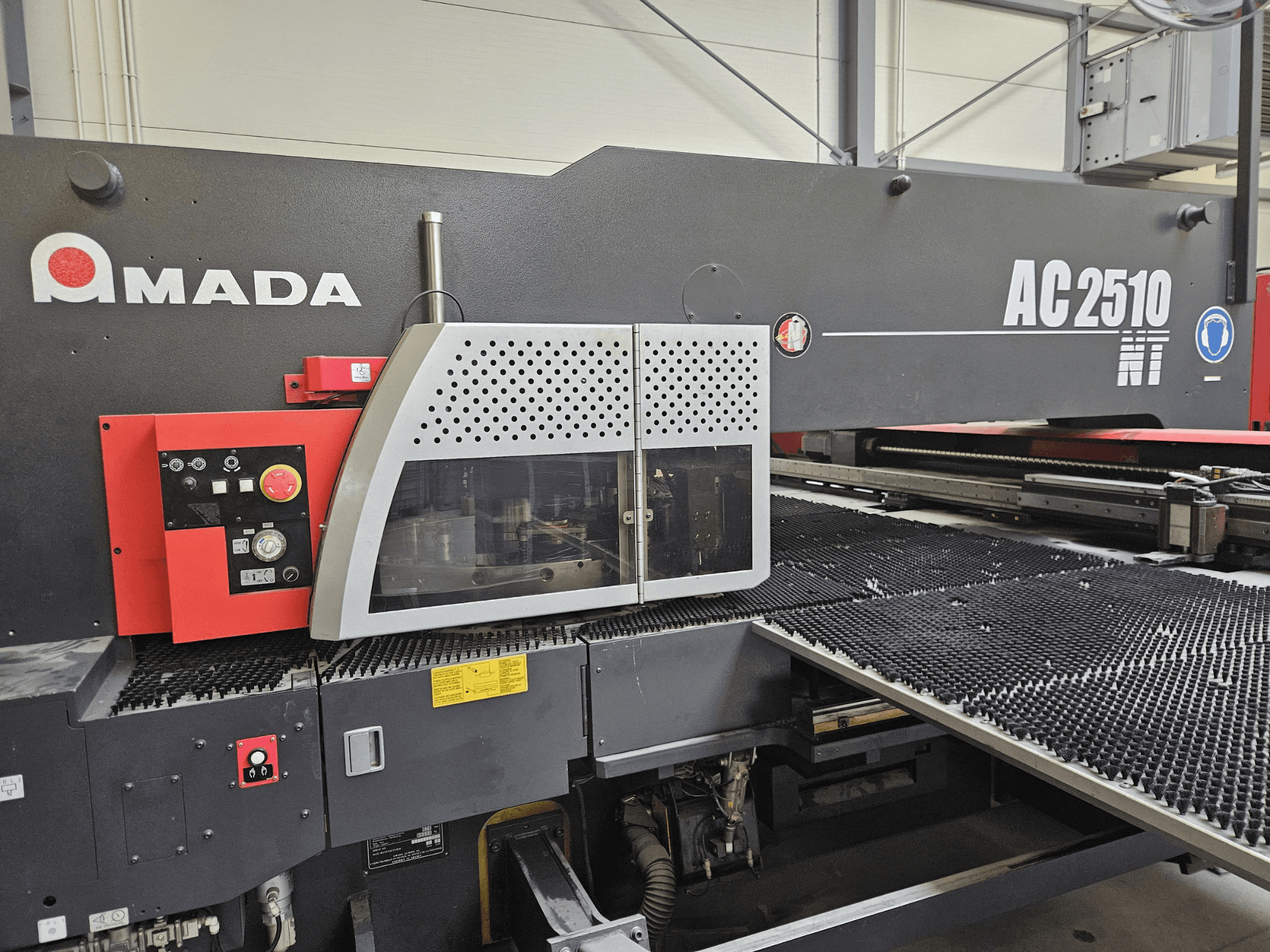 "Amada AC2510 NT" lazerinio pjaustymo staklės, vaizdas iš priekio, valdymo skydelis su rankenėlėmis, mygtukais ir apsauginiu dangteliu.