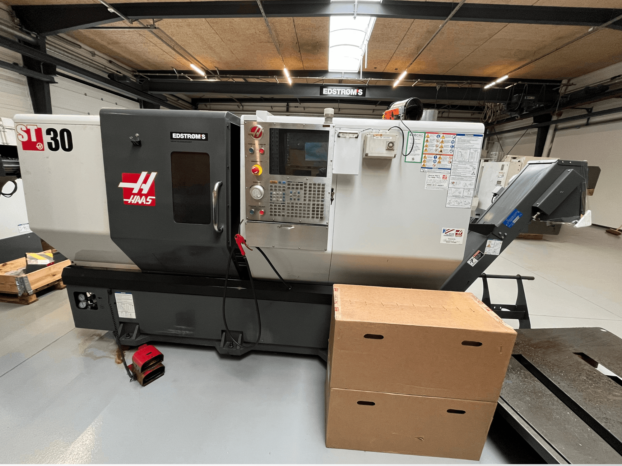 Haas ST-30 CNC staklės, žiūrint iš šono, su valdymo skydeliu, modelio ženkliuku ir dviem kartoninėmis dėžėmis šalia.