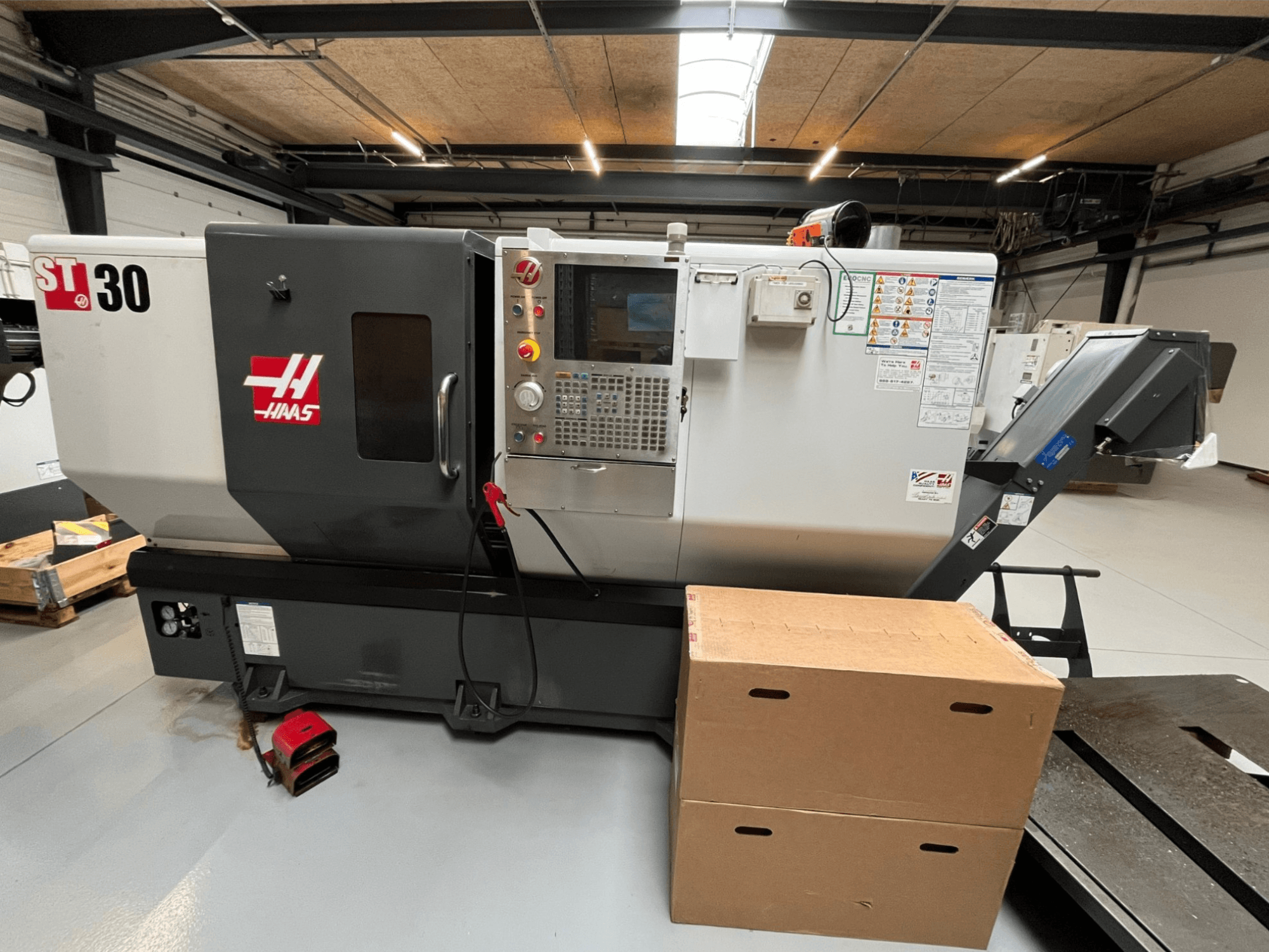 Haas ST-30 CNC tekinimo staklės, vaizdas iš priekio, valdymo skydelis su mygtukais, avarinis stabdys ir dvi kartoninės dėžės.