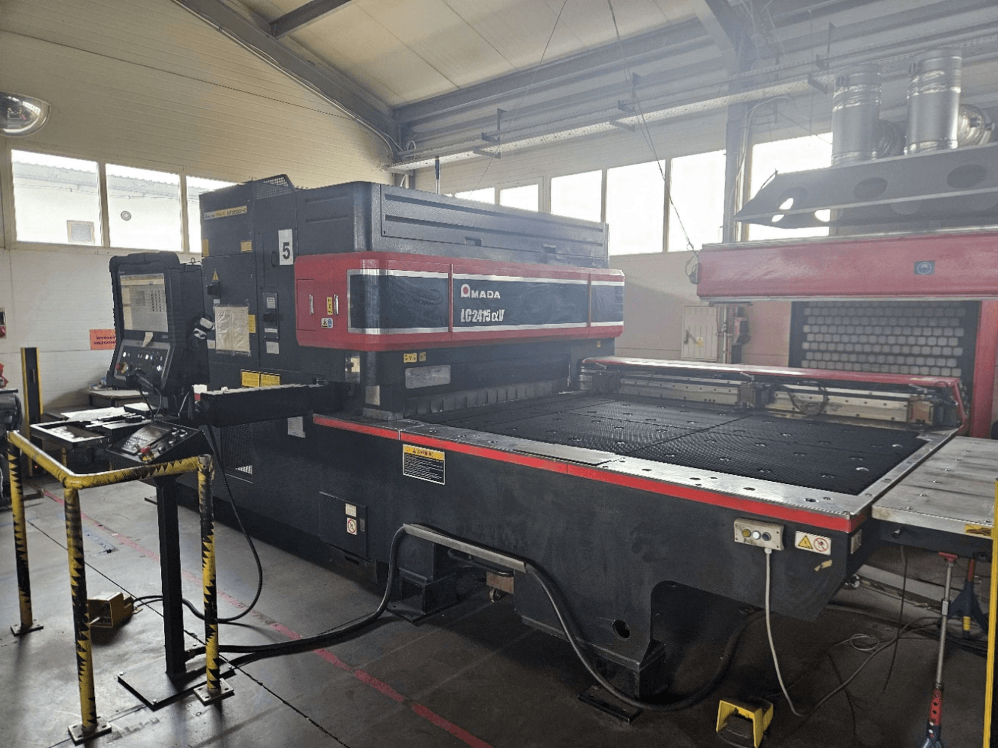 AMADA LC2415 Alpha 5 CO₂ lazerio pjovimo staklės, vaizdas iš priekio, kuriame matomas valdymo skydas ir pjovimo stalas su apsauginėmis užtvaromis.