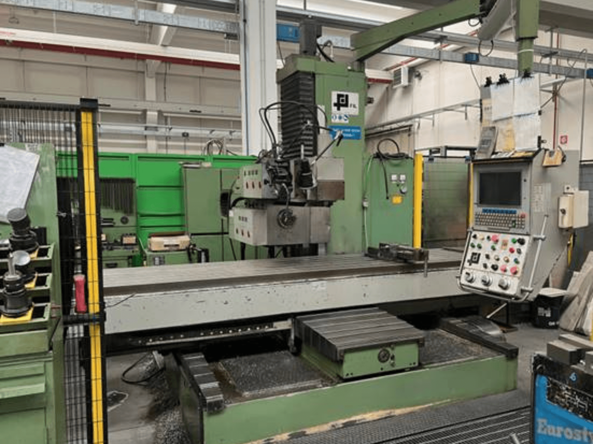 JFL pramoninės frezavimo staklės, vaizdas iš priekio su valdymo pultu, dideliu plokščiu stalu ir metalo drožlėmis ant grindų.