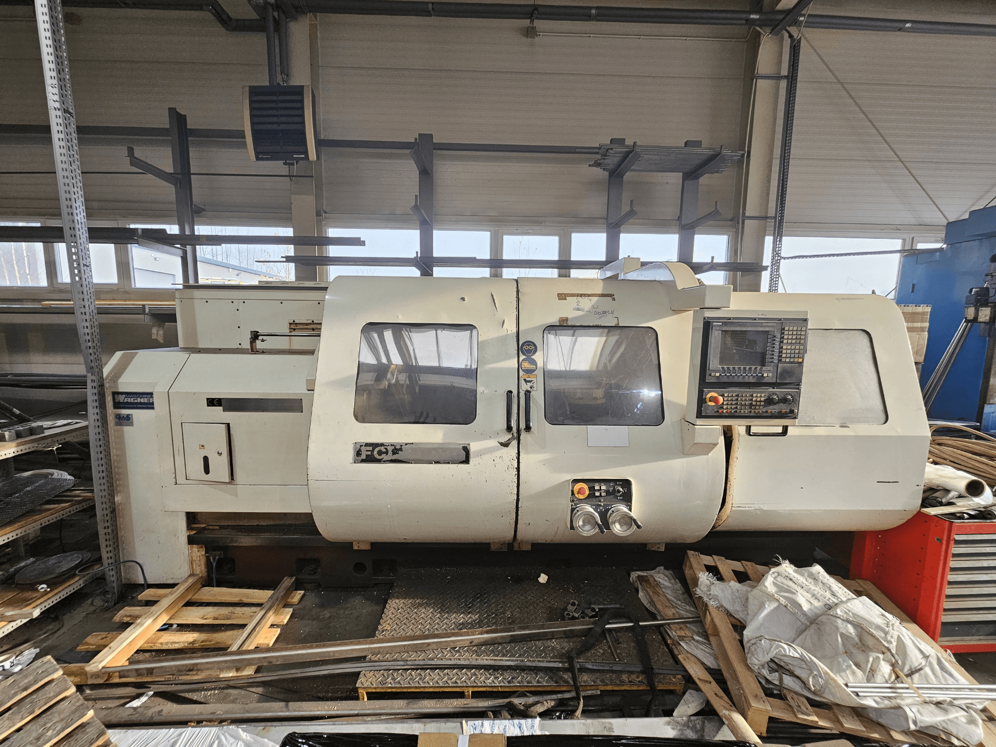 Wabeco CNC staklės, vaizdas iš šono, valdymo skydelis, langai ir darbo vieta; mašinos ant medinių padėklų dirbtuvėse.