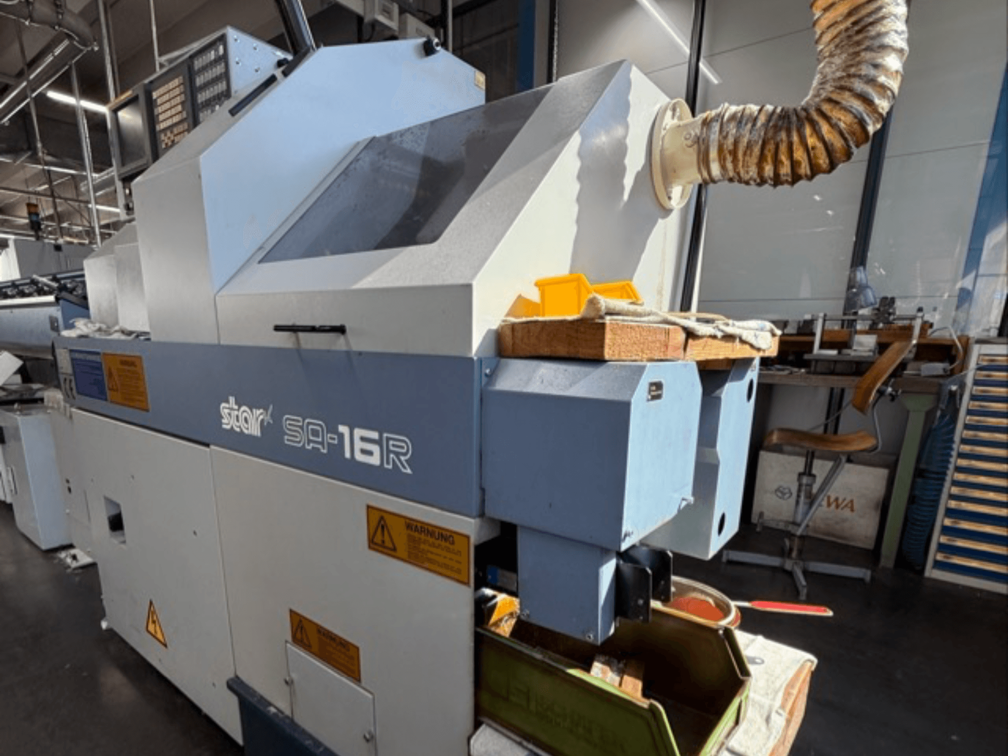 "Stark SA-16R" staklės, žiūrint iš priekio, su kūginiu išmetimo vamzdžiu ir darbo vieta su geltonais įrankiais ir įspėjamaisiais užrašais.