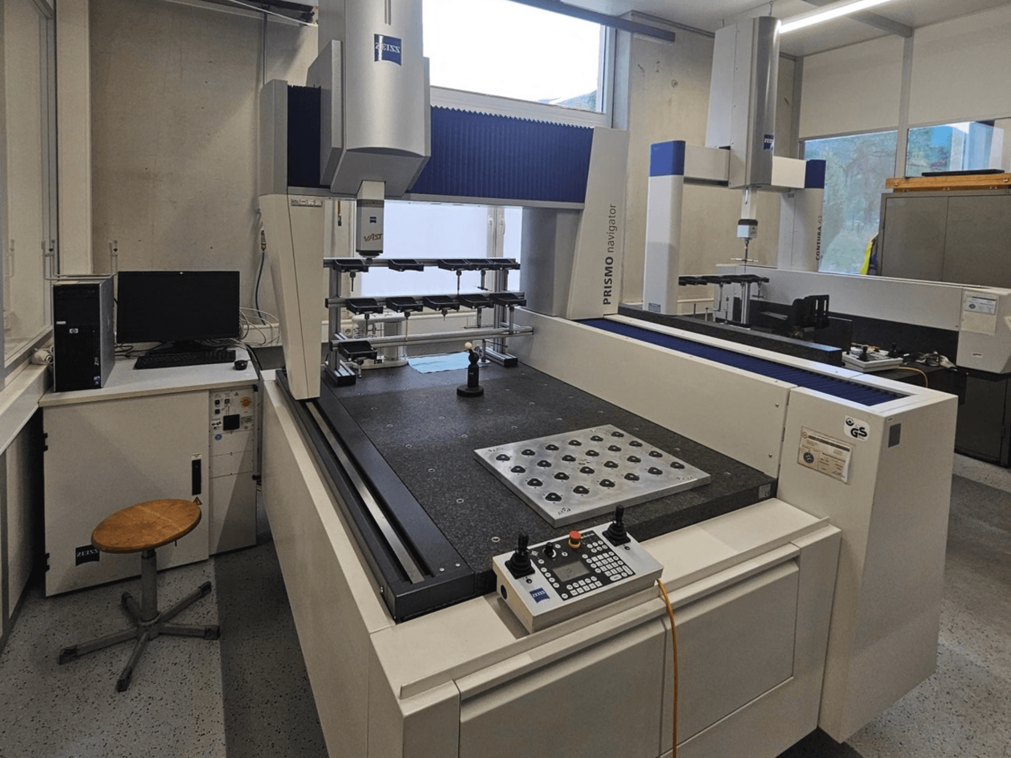 ZEISS Prismo Navigator 7 CMM laboratorijoje, vaizdas iš priekio, kuriame matomas valdymo skydas, darbo stalas ir monitorius.