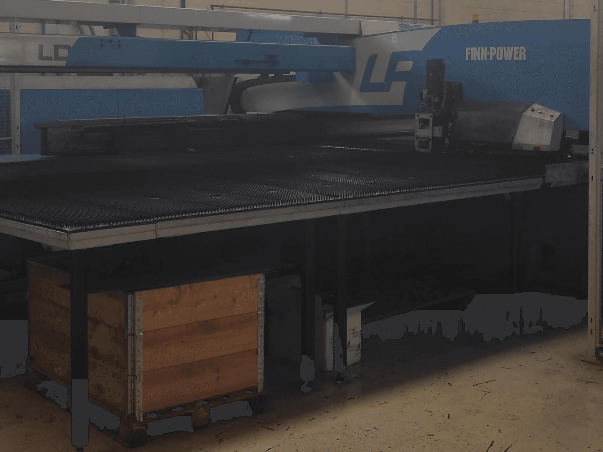 "Finn-Power LP" pjovimo mašina gamykloje, vaizdas iš priekio su tekstūruotu metaliniu stalu ir medine dėže po juo.