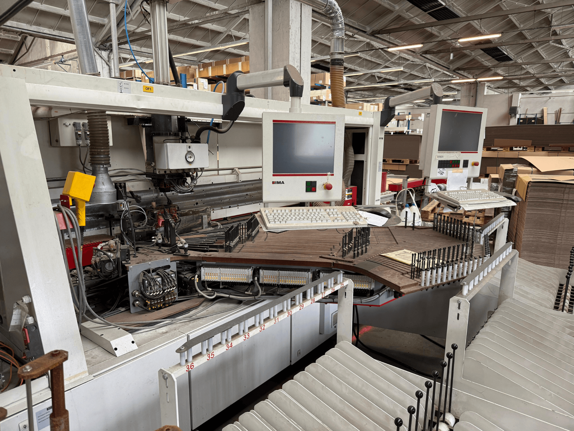 IMA tiksliojo medžio apdirbimo staklės su dviem monitoriais ir valdymo skydeliais, vaizdas iš priekio, rodantis konvejerį ir įrankių zoną.