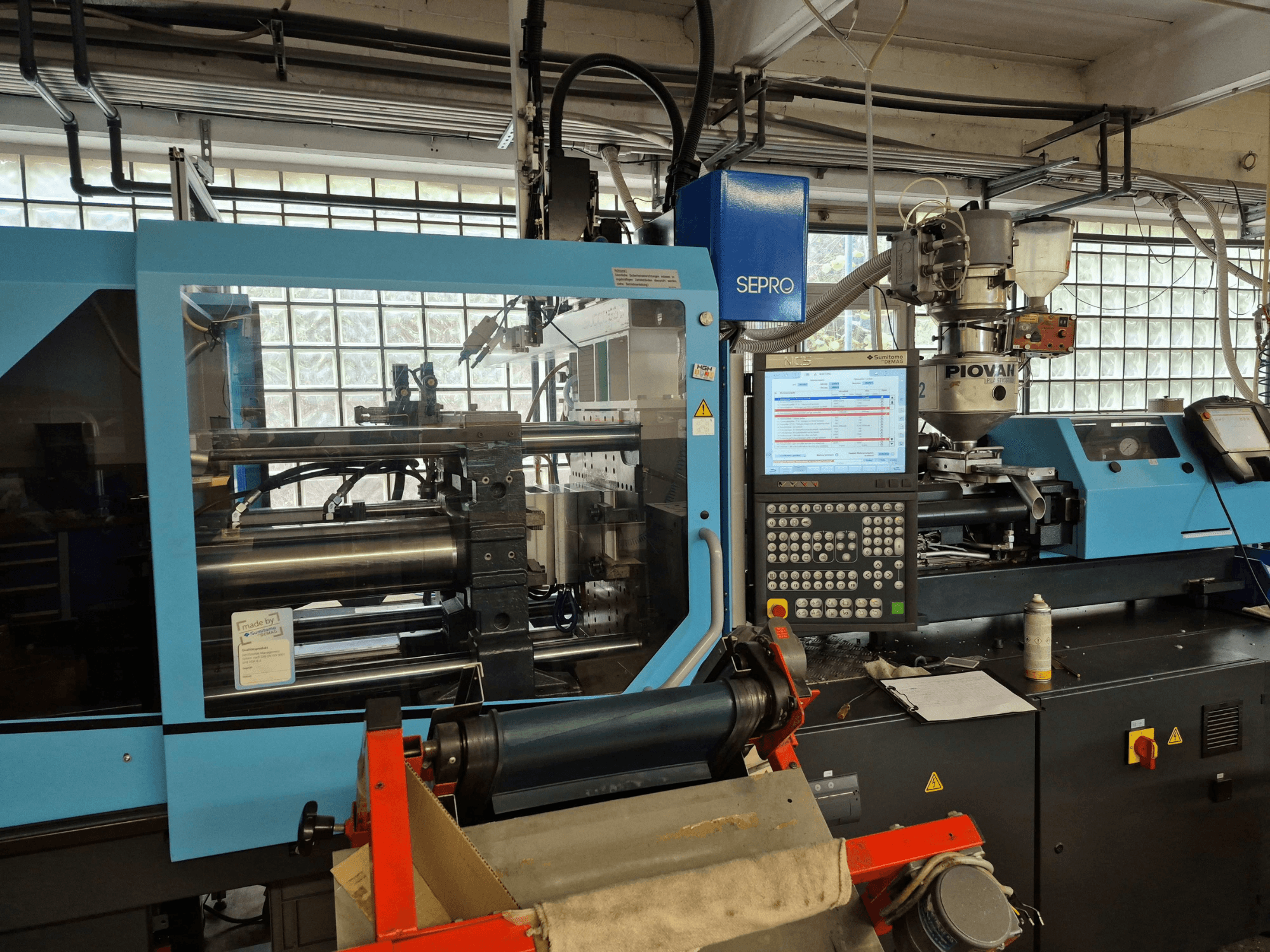 Hidraulinė liejimo mašina „SUMITOMO SHI DEMAG Systec 120/470-430“, vaizdas iš priekio, kuriame matomas valdymo pultas ir padavimo sistema.
