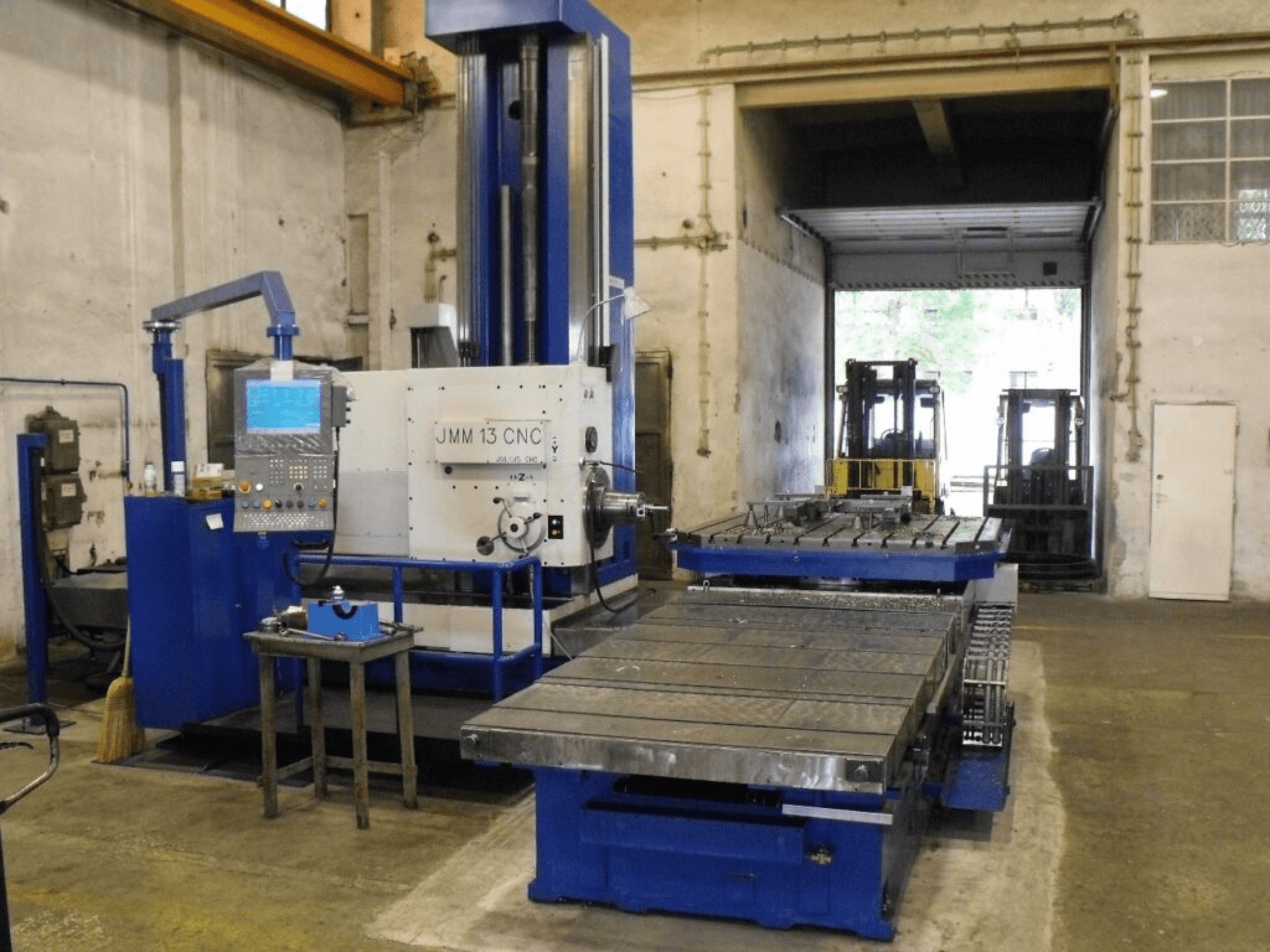 JMM 13 CNC frezavimo staklės dirbtuvėse, vaizdas iš priekio, su valdymo pultu, dideliu darbo stalu ir pramoniniu fonu.