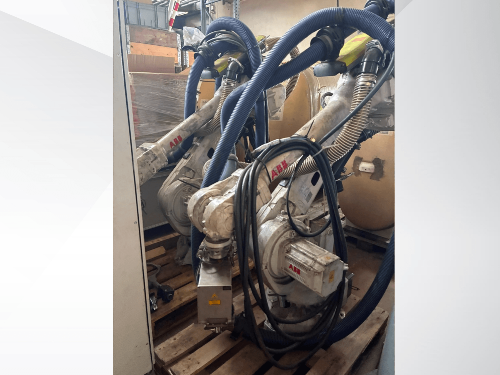 ABB IRB 4600-40/2.55 robotas, vaizdas iš šono, kuriame matomos dvi rankos ir prie jų pritvirtintos žarnos; robotas sumontuotas ant padėklų sandėlio aplinkoje.
