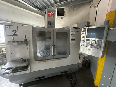 Haas VF-2 CNC staklės, vaizdas iš priekio, su valdikliais, įrankių laikikliu ir permatomomis durelėmis su matomais įrankiais spintelės viduje.