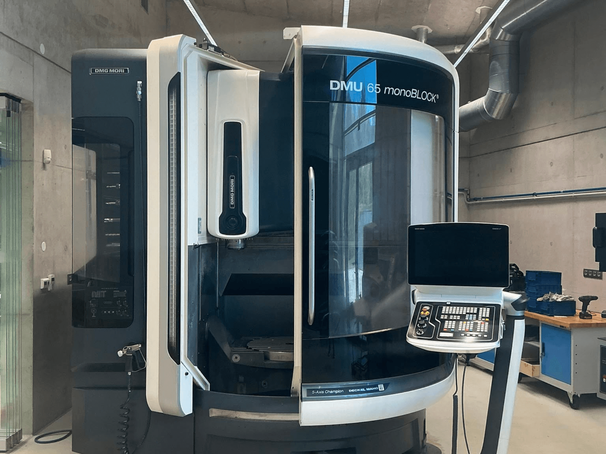 DMG MORI DMU 65 monoBLOCK vertikalus apdirbimo centras, vaizdas iš priekio, kuriame matomas valdymo pultas ir staklių detalės dirbtuvės aplinkoje.