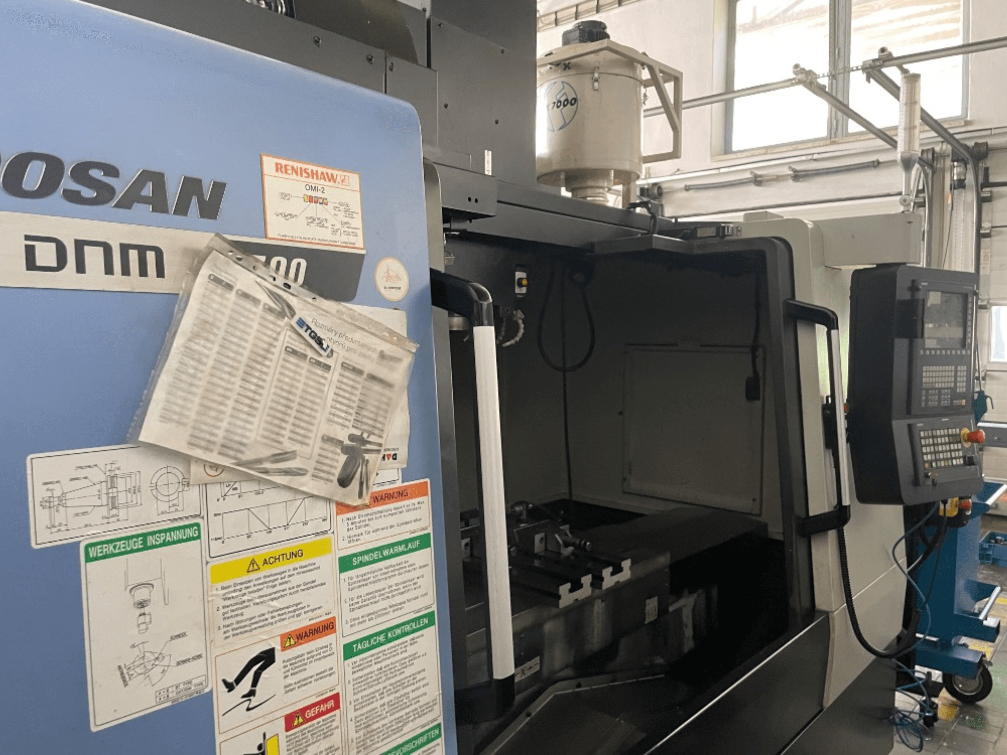 DOOSAN DNM 5700 vertikalusis apdirbimo centras, priekinis vaizdas, rodomas valdymo skydelis, saugos etiketės ir staklių detalės.