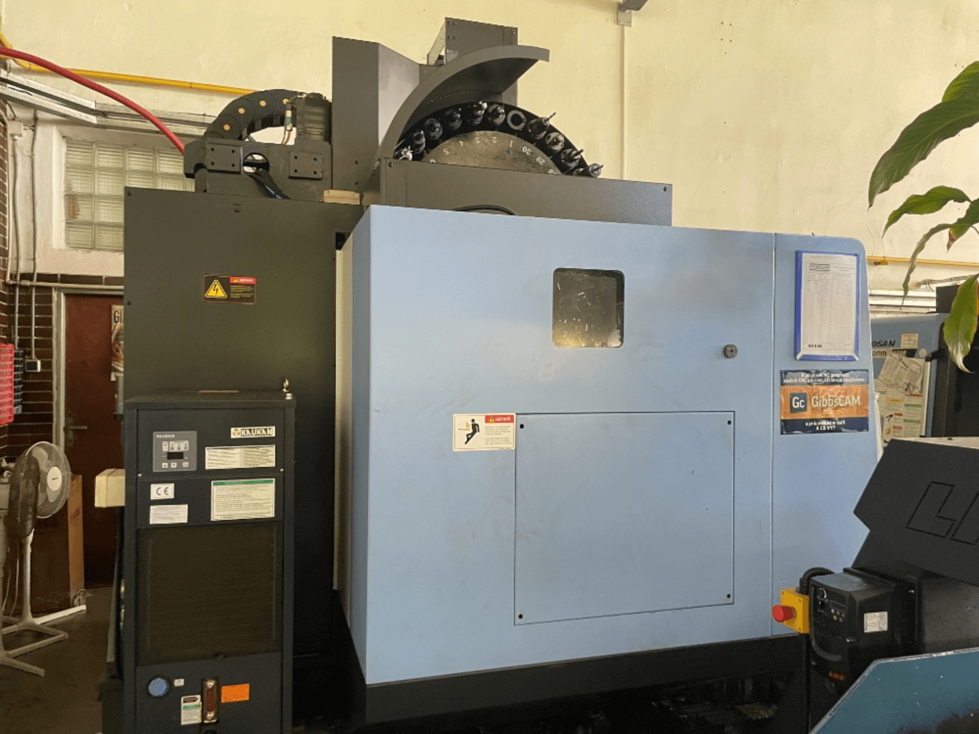 Vertikalaus apdirbimo centro DOOSAN DNM 5700 priekinis vaizdas, kuriame matomas valdymo skydelis, saugos etiketės ir staklių funkcijos.