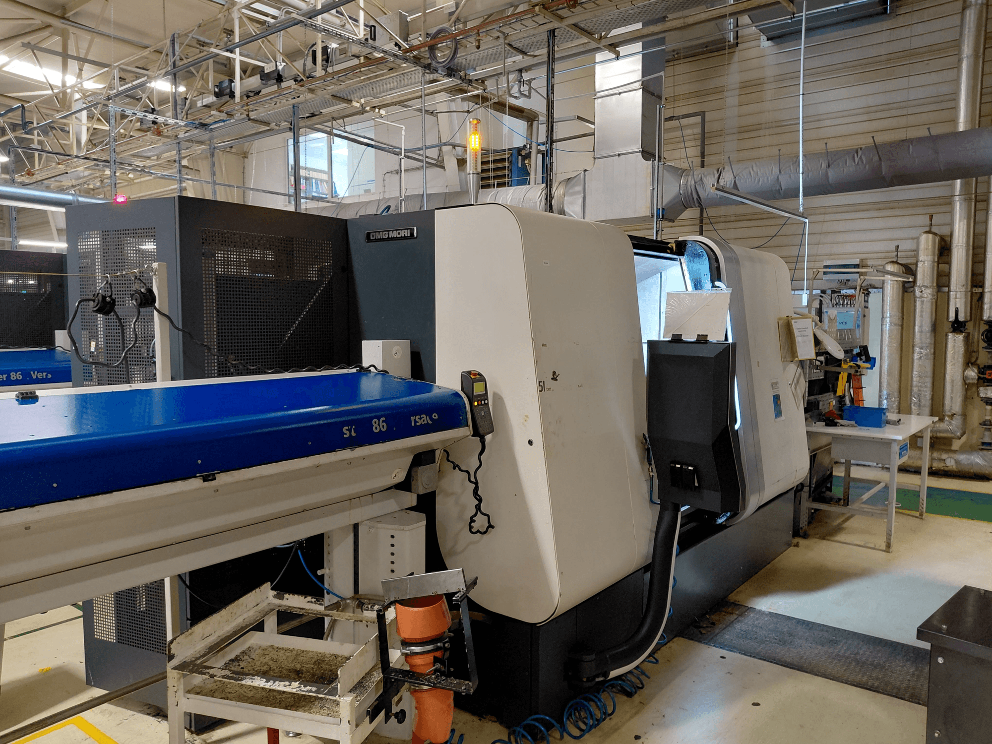 DMG MORI Sprint 65 linijinės daugiasvirtės tekinimo staklės, šoninis vaizdas su valdymo pultu ir konvejeriu, matomu dirbtuvėse.