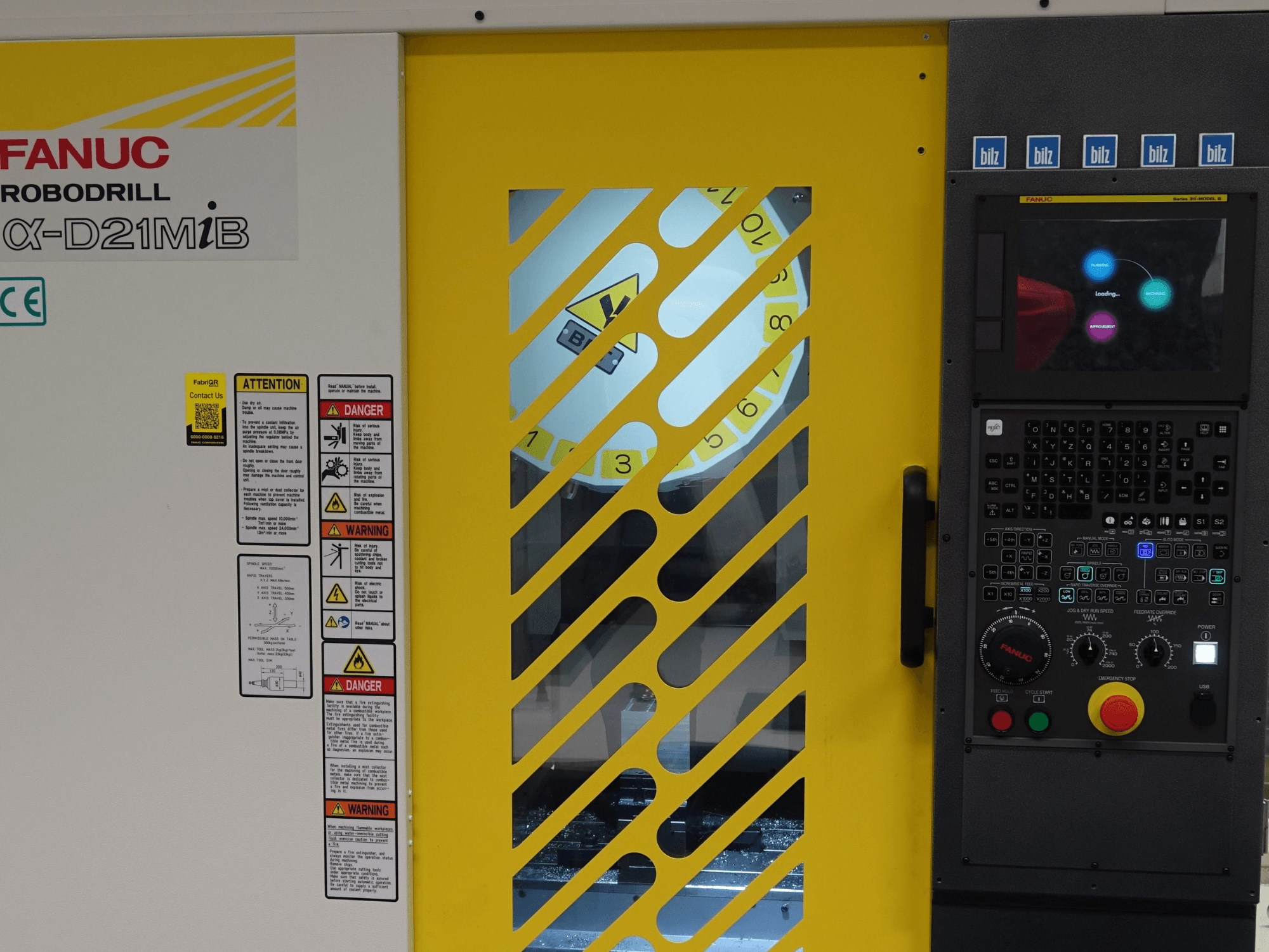 FANUC α-D21MiB vertikalaus apdirbimo centro vaizdas iš priekio, kuriame matomi saugos įspėjimai, valdymo pultas ir geltonos spalvos saugos durys.