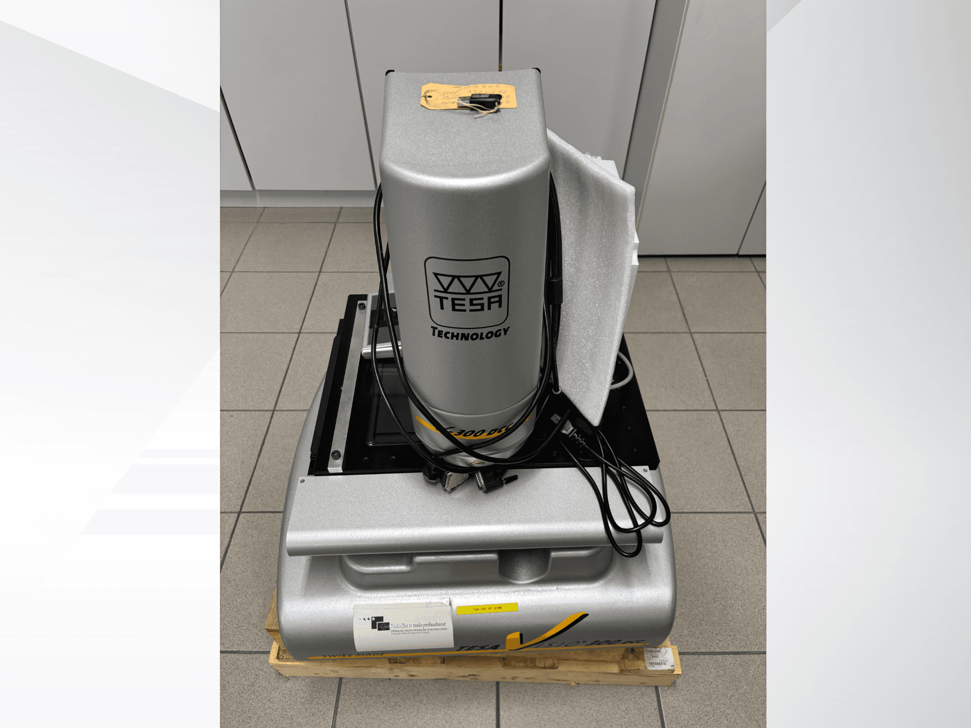 TESA Visio 300 DCC optinis matavimo aparatas iš priekio su kabeliais ant medinio padėklo ir matoma apsauginė pakuotė.