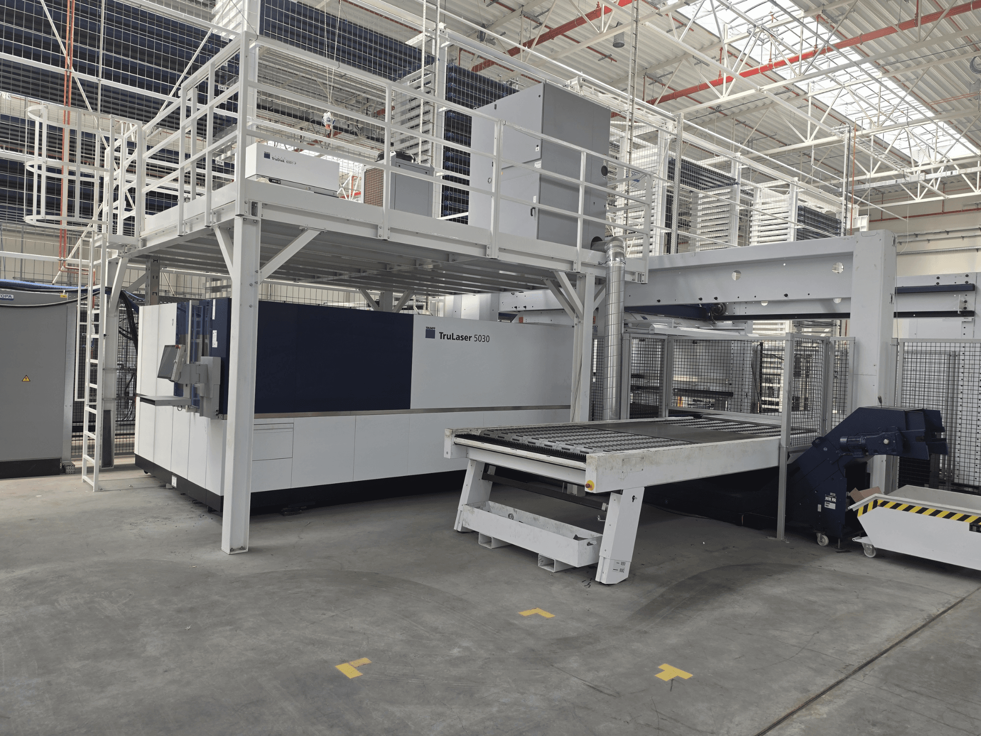 „TRUMPF TruLaser 5030 fiber L76 FMC“ staklės dirbtuvėse, vaizdas iš priekio, kuriame matoma pjovimo zona ir pakrovimo bei iškrovimo mechanizmas.