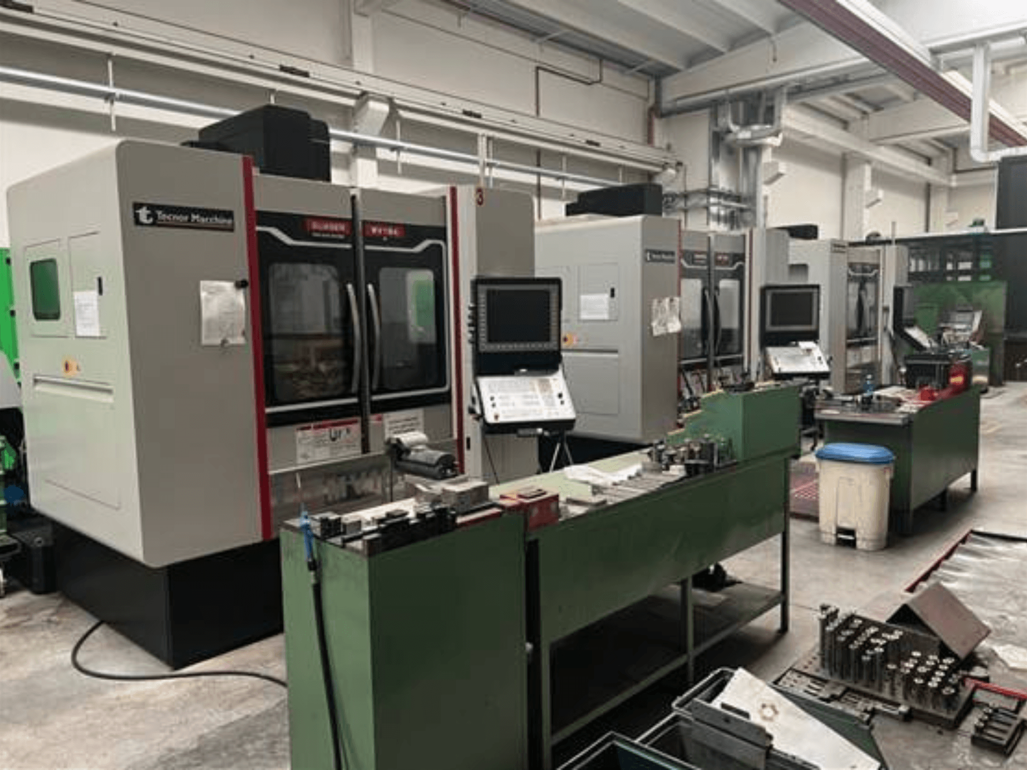 Keturios "Tecnoer Marchine" CNC staklės dirbtuvėse, vaizdas iš priekio, su įrankiais ir dalimis ant žalių darbo stalų.