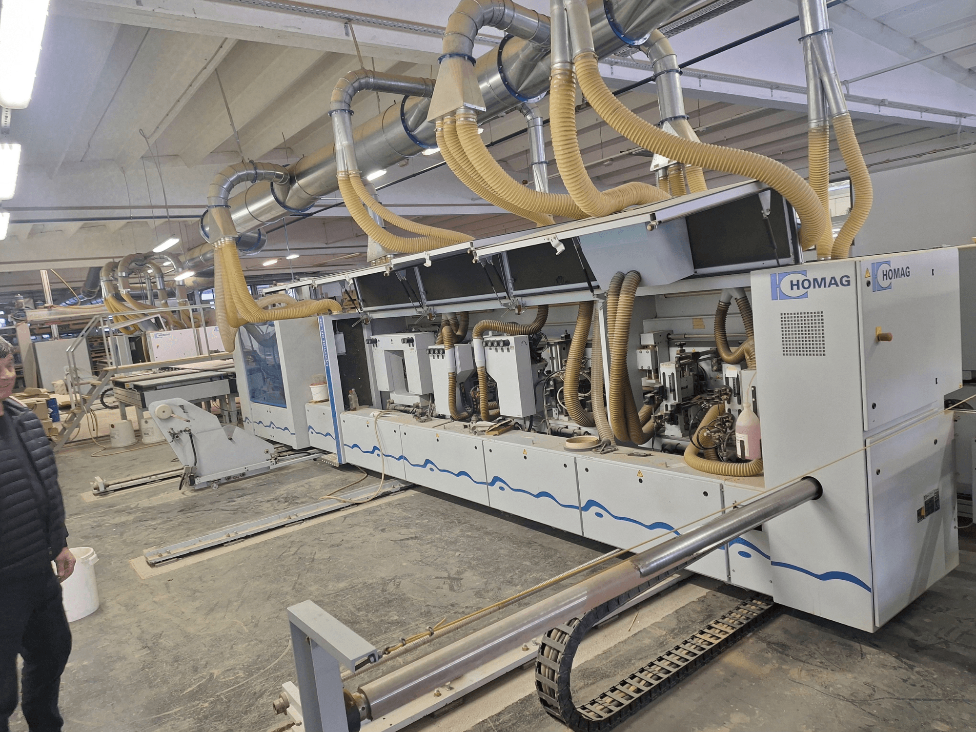 HOMAG medžio apdirbimo staklės gamykloje, vaizdas iš priekio, kuriame matomi keli velenai ir dulkių nusiurbimo sistema.