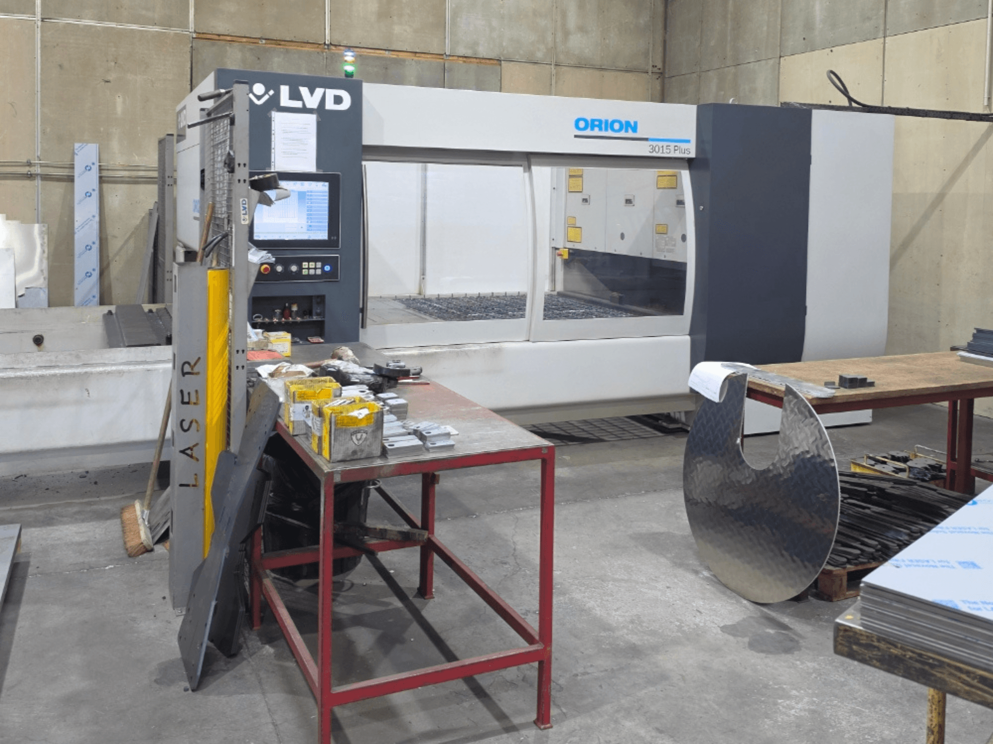 LVD „Orion 3015 Plus“ CO₂ lazerio pjovimo staklės, vaizdas iš priekio su valdymo pultu; ant darbo stalo, apsuptos metalinių detalių.