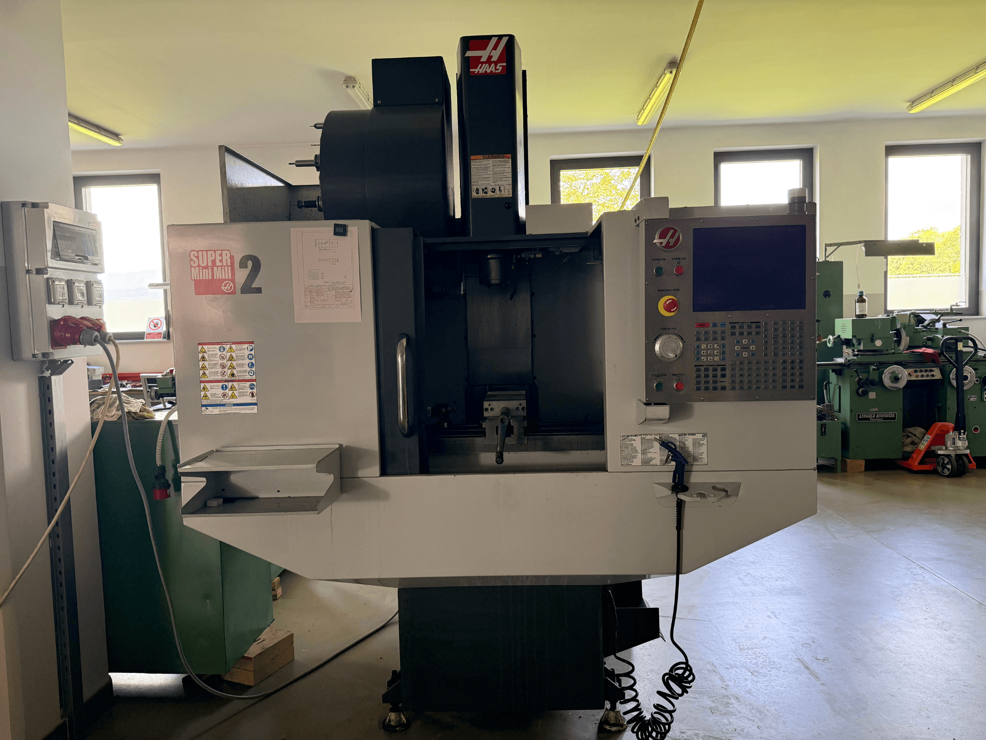 Vertikalaus apdirbimo centro "Haas Mini Mill 2" vaizdas iš priekio - valdymo skydelis ir įrankių zona su įspėjamaisiais užrašais.