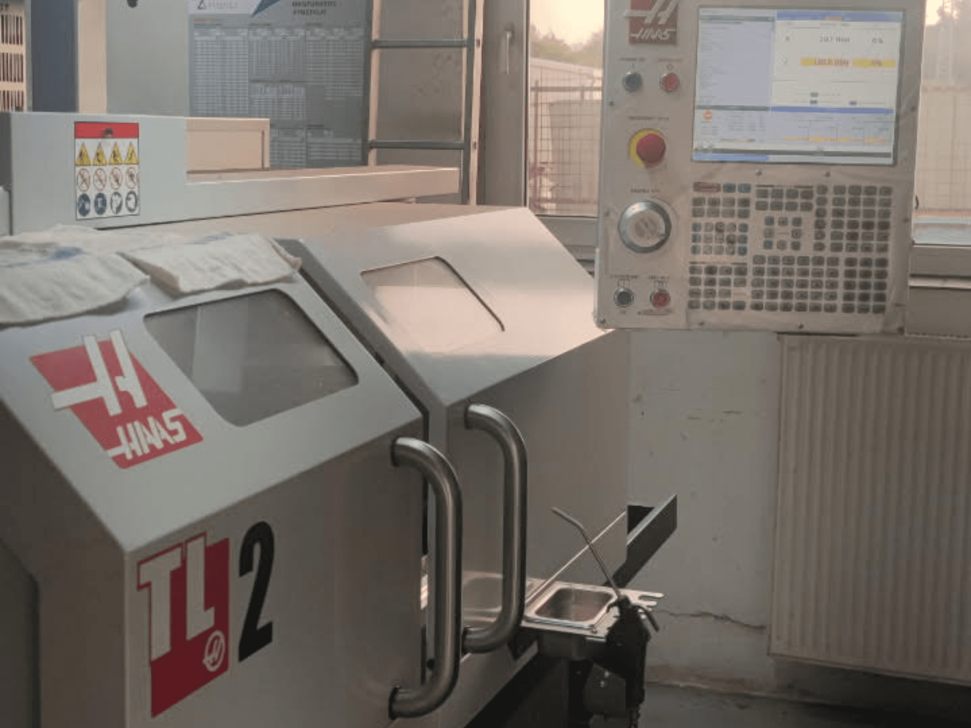 Horizontaliojo tekinimo staklių "Haas TL-2" priekinis vaizdas, kuriame matyti valdymo skydelis, metalinis korpusas ir saugos ženklai.