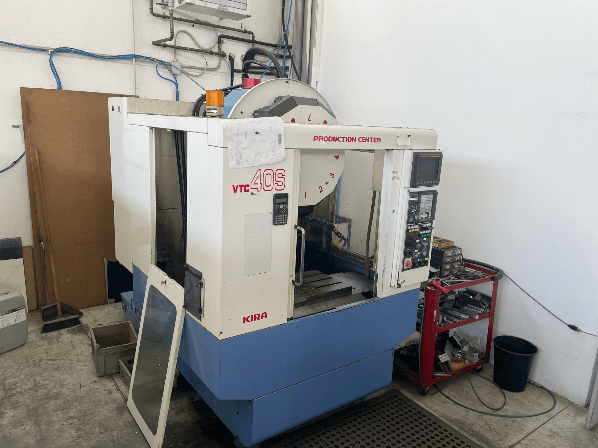 KIRA VTC 40S mechaninio apdirbimo centras iš šono, su valdymo skydeliu, atidarytomis durelėmis ir įrankiais ant šalia esančio sandėliavimo vežimėlio.