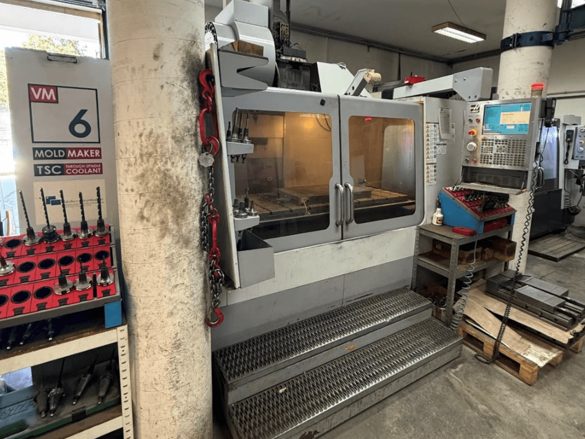 HAAS VM-6 HE vertikalus apdirbimo centras, vaizdas iš priekio, kuriame matomi valdymo elementai, aušinimo sistema ir įrankių saugykla.