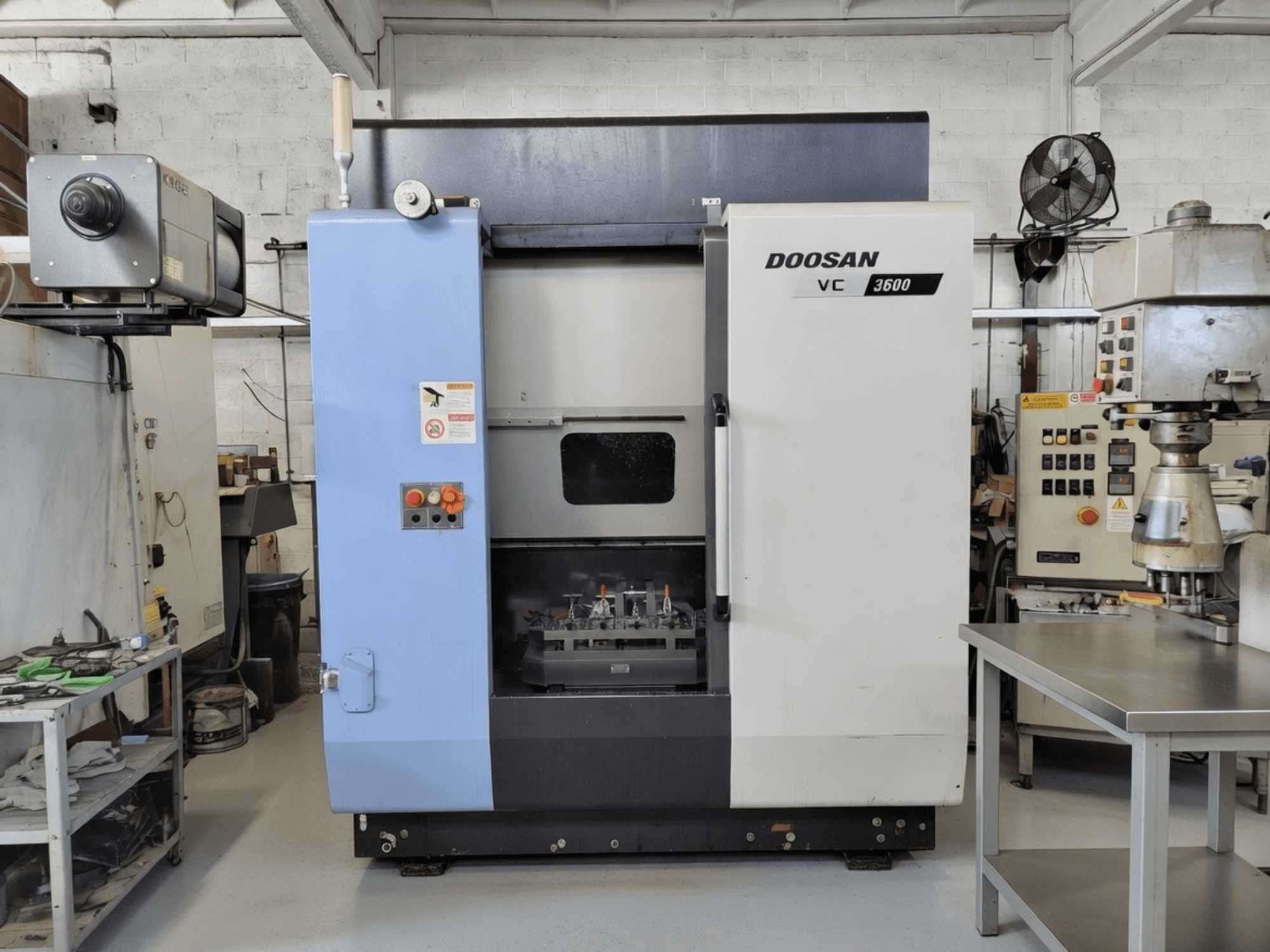 DOOSAN VC 3600 vertikalusis apdirbimo centras iš priekio, su valdymo pultu ir atviromis durimis, pro kurias matoma darbo vieta.
