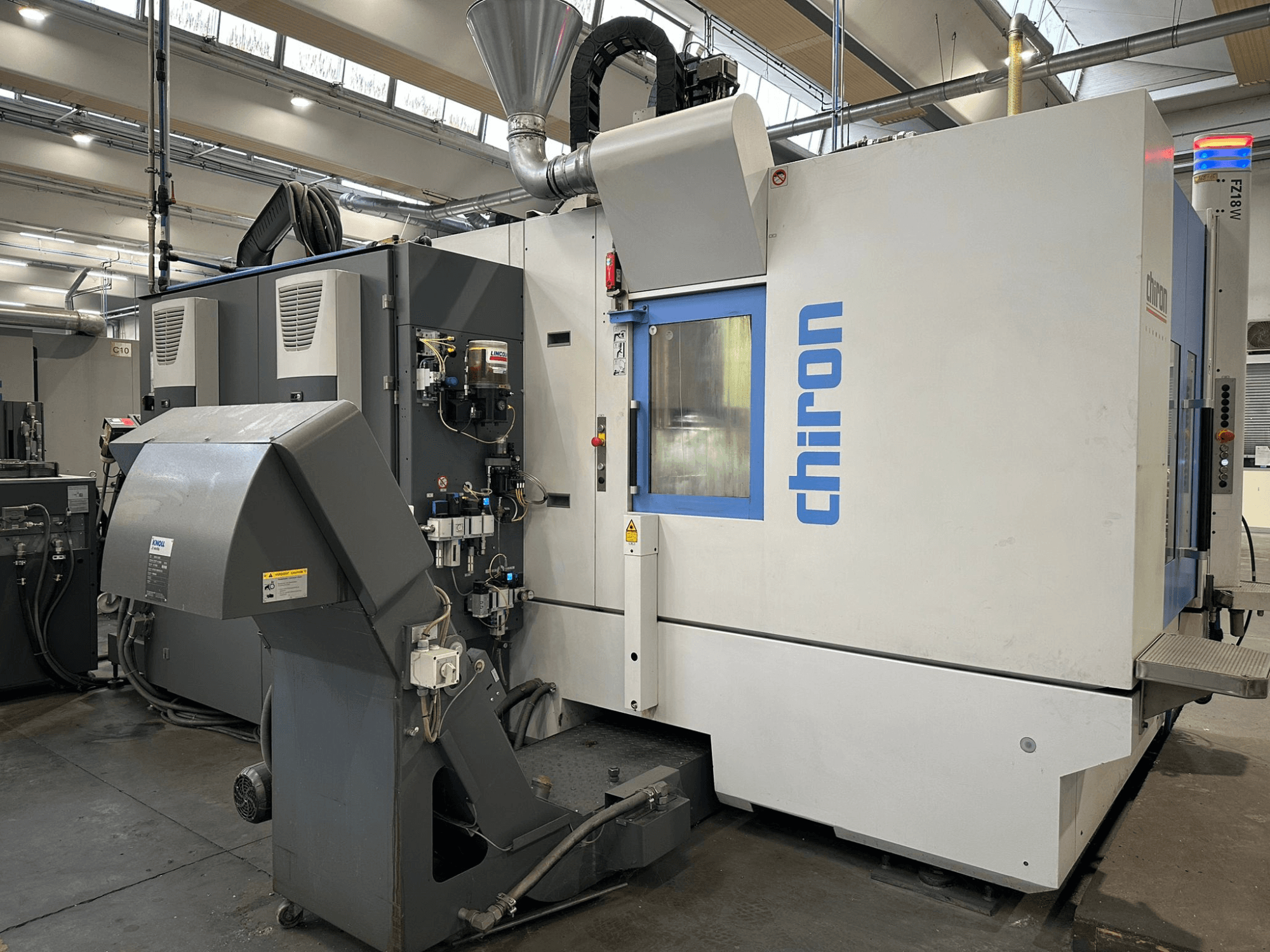 Pilkos ir baltos spalvos "Chiron CNC" staklės, vaizdas iš priekio, su valdikliais, vamzdžiais ir dulkių ištraukimo gaubtu.