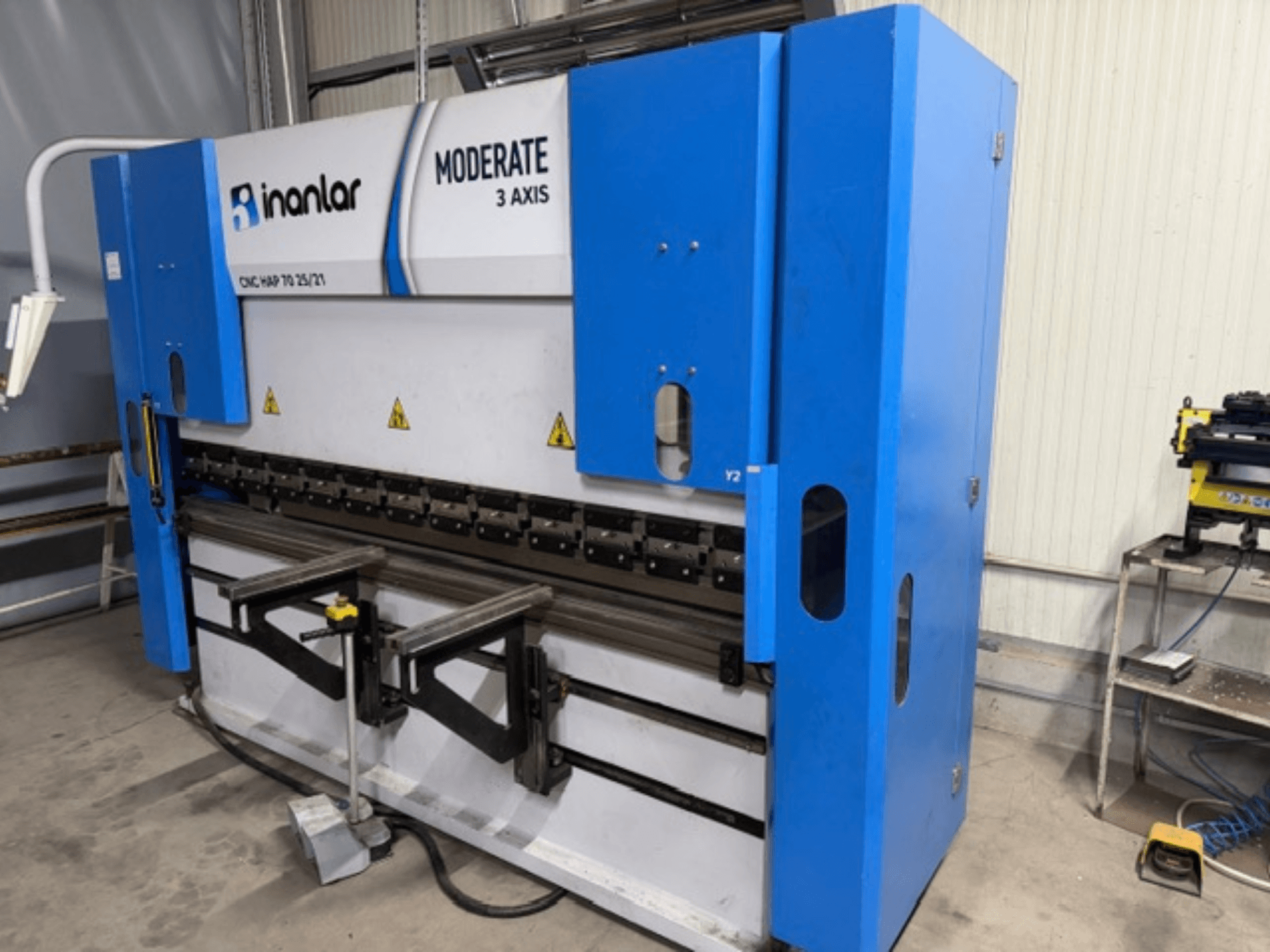 Mėlynos spalvos "Inanlar Moderate" 3 ašių CNC staklės, vaizdas iš priekio, ant korpuso matomos valdymo svirtys ir saugos etiketės.