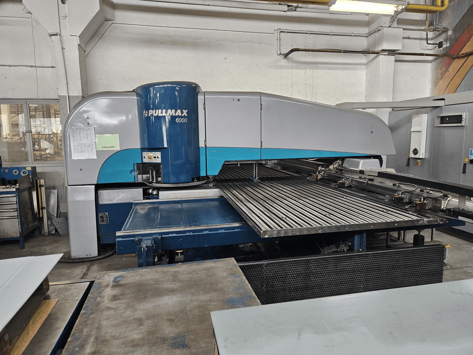 "Pullmax 6000" CNC perforavimo staklių "Pullmax 6000" vaizdas iš priekio, demonstruojantis didelį plokščią stalviršį ir metalinį karkasą dirbtuvėse.