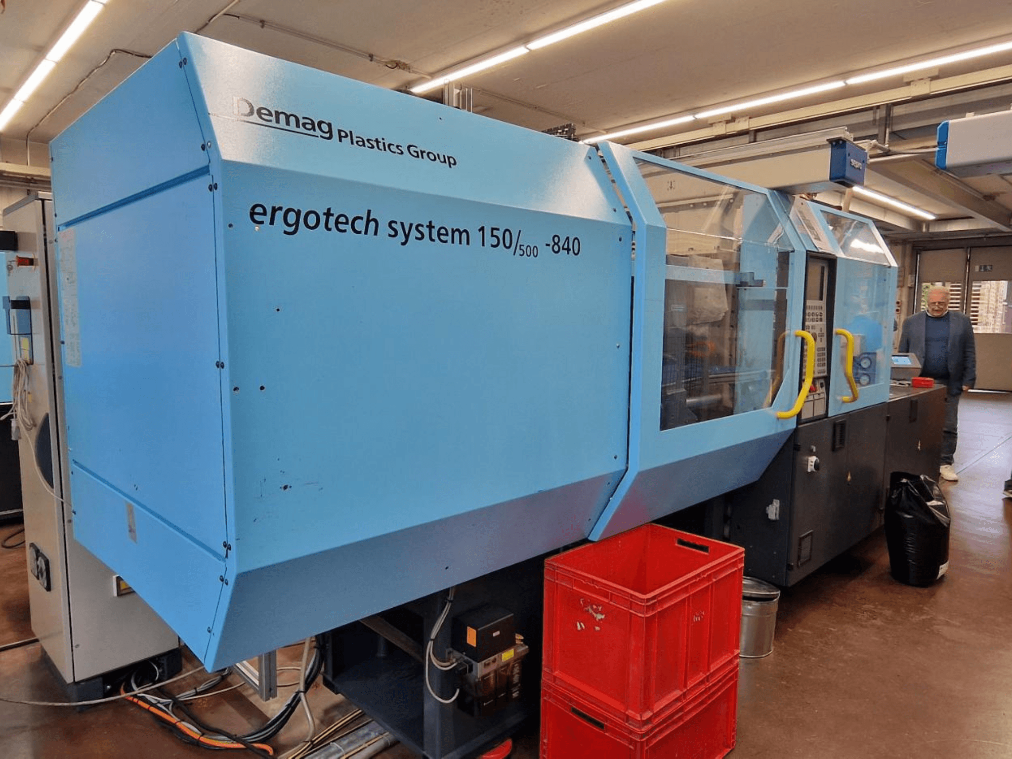 Mėlynos spalvos "Demag Ergotech System 150/500-840" šoninis vaizdas su dėžėmis ir operatoriumi fone.