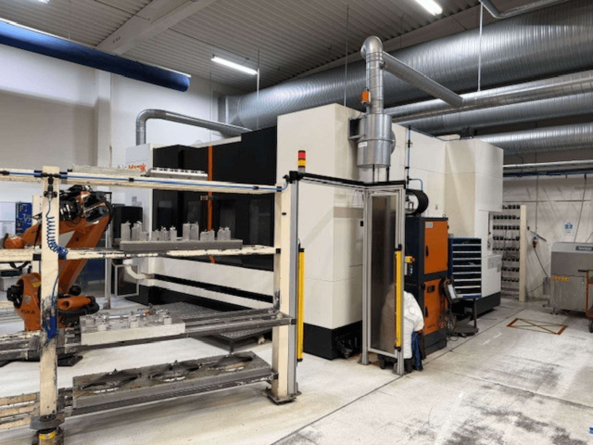 Pramoninės CNC staklės su robotine ranka ir apsauginėmis užtvaromis, kuriose matomas priekinis įrenginio vaizdas erdvioje darbo vietoje.