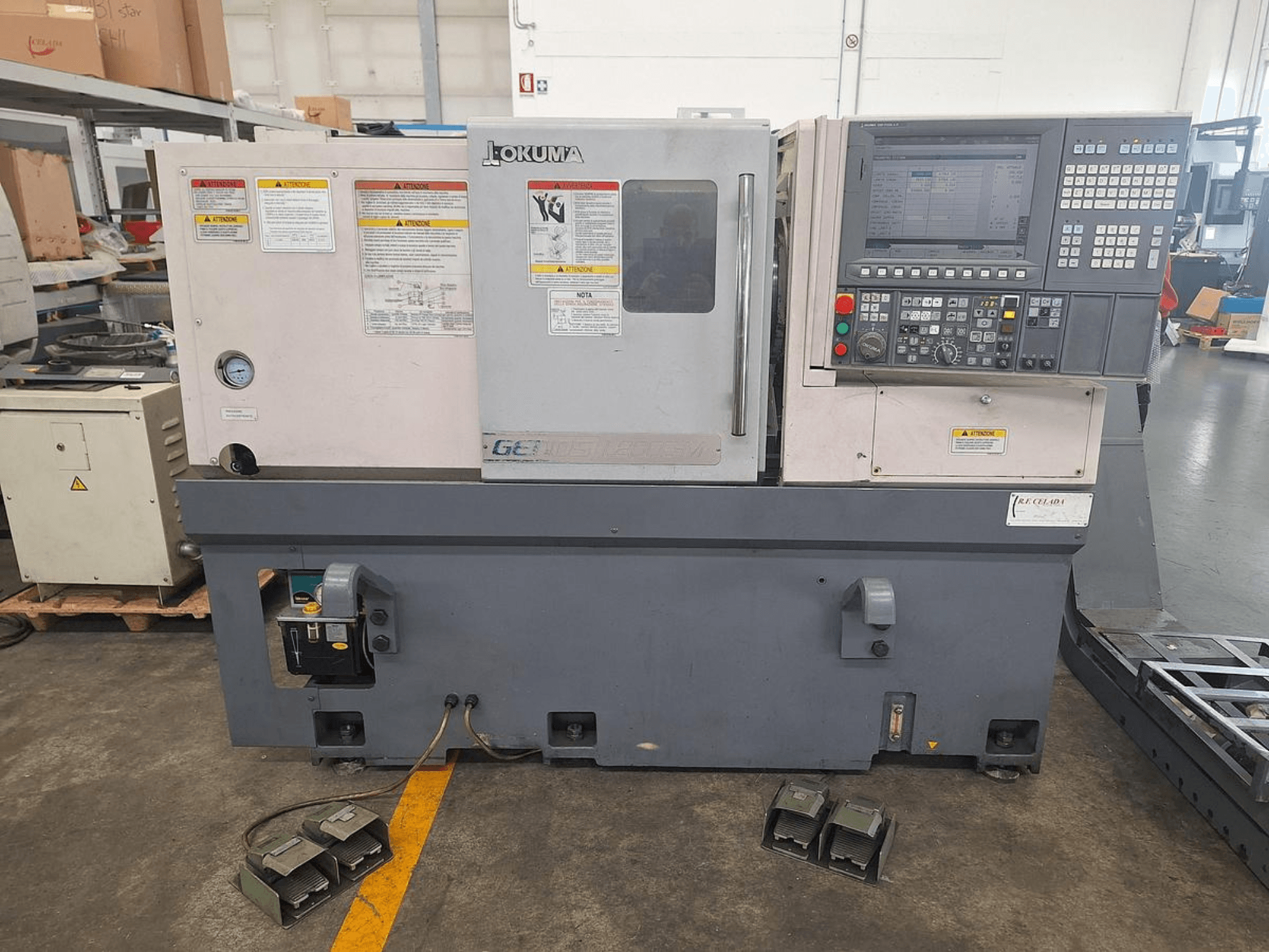 Okuma GENOS L200E-M horizontaliosios tekinimo staklės iš šono; valdymo skydelis, saugos įspėjimai ir kojiniai pedalai.