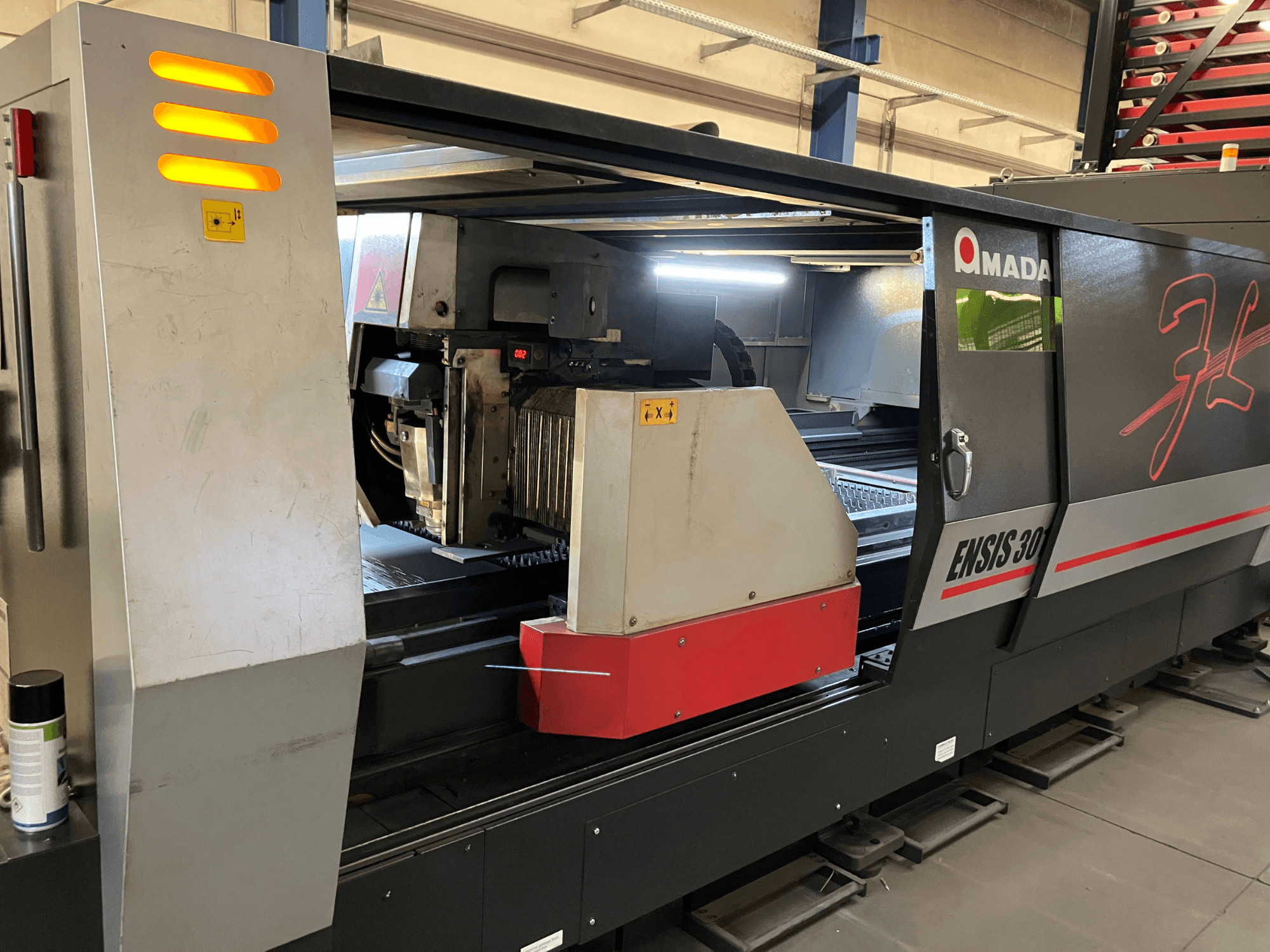 AMADA ENSIS 3015 AJ pluoštinio lazerio pjovimo staklės, vaizdas iš priekio, pjovimo galvutė ir valdymo įtaisai metaliniame rėmelyje.