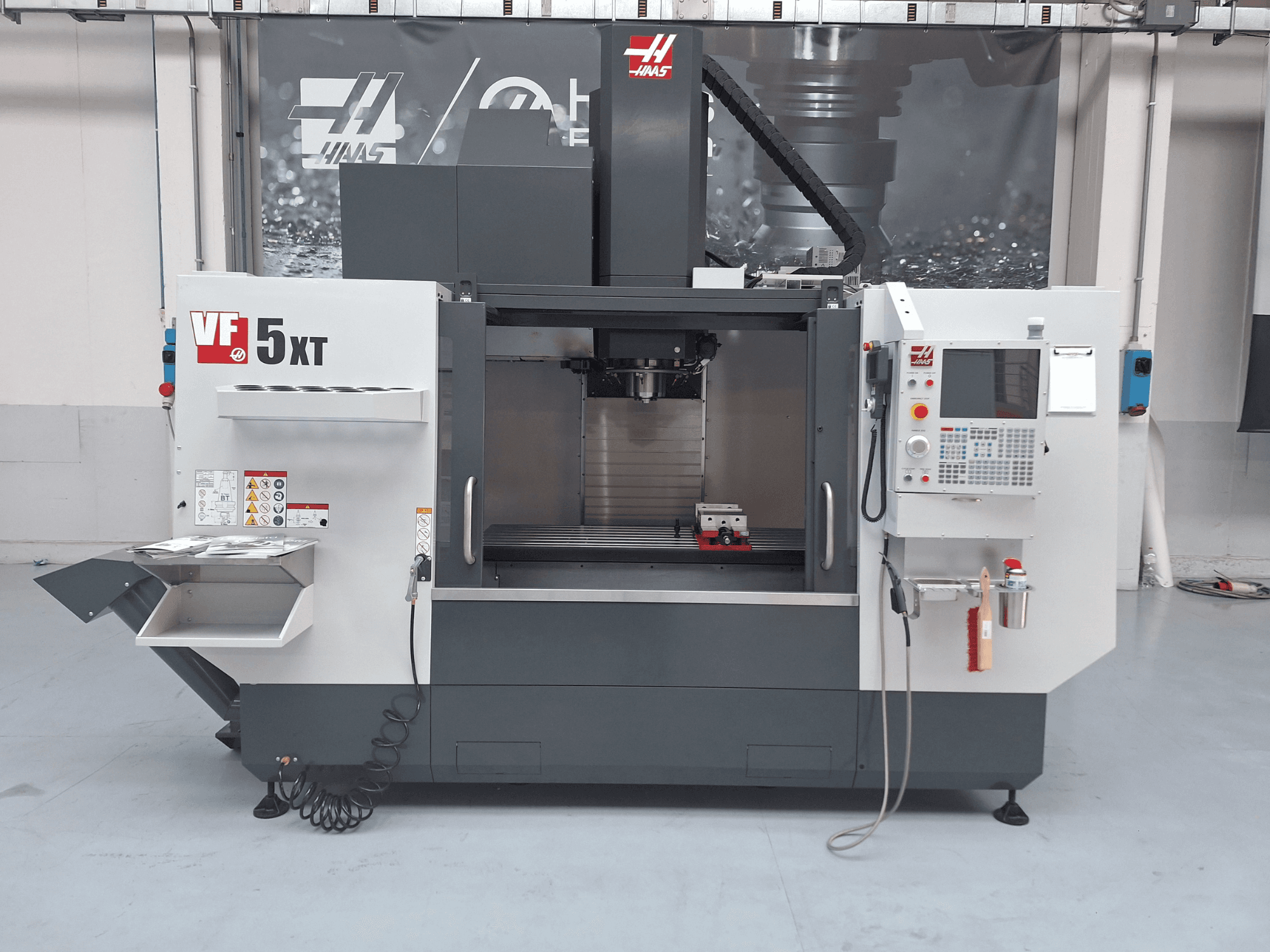 Vertikalusis apdirbimo centras „HAAS VF-5/50XT“, vaizdas iš priekio, kuriame matomas valdymo pultas, suklio zona ir įrankių laikymo vieta.