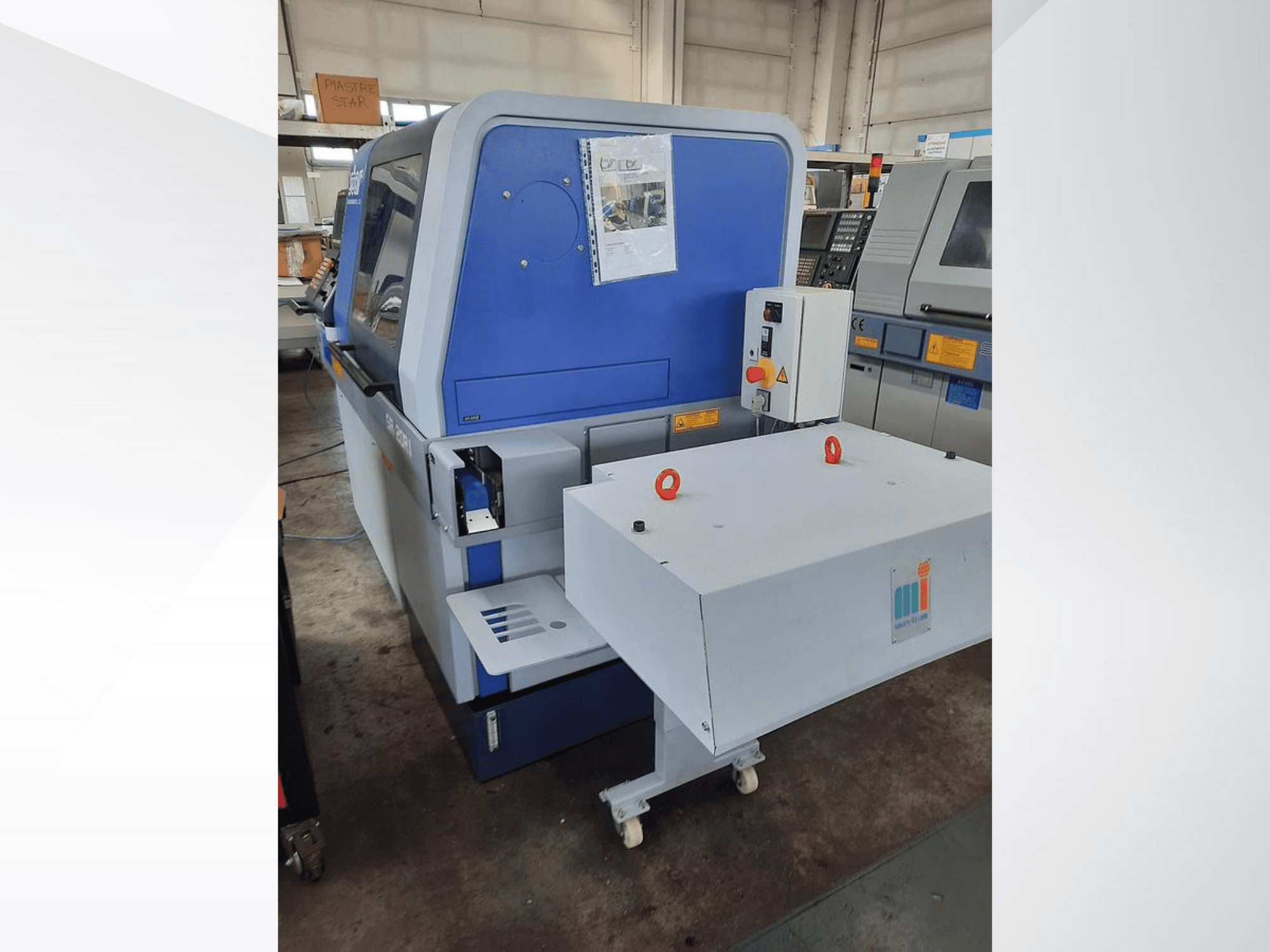 Mėlyna pramoninė mašina su baltu konvejeriu, kurioje matyti valdymo skydelis ir pritvirtintos saugos priemonės, vaizdas iš šono.