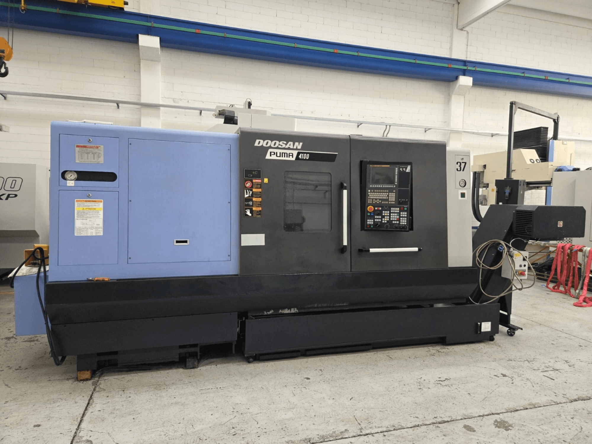 "Doosan Puma 4100 CNC" tekinimo staklės, vaizdas iš priekio; valdymo skydelis, mėlynas maitinimo blokas ir išsamios darbui skirtos etiketės.