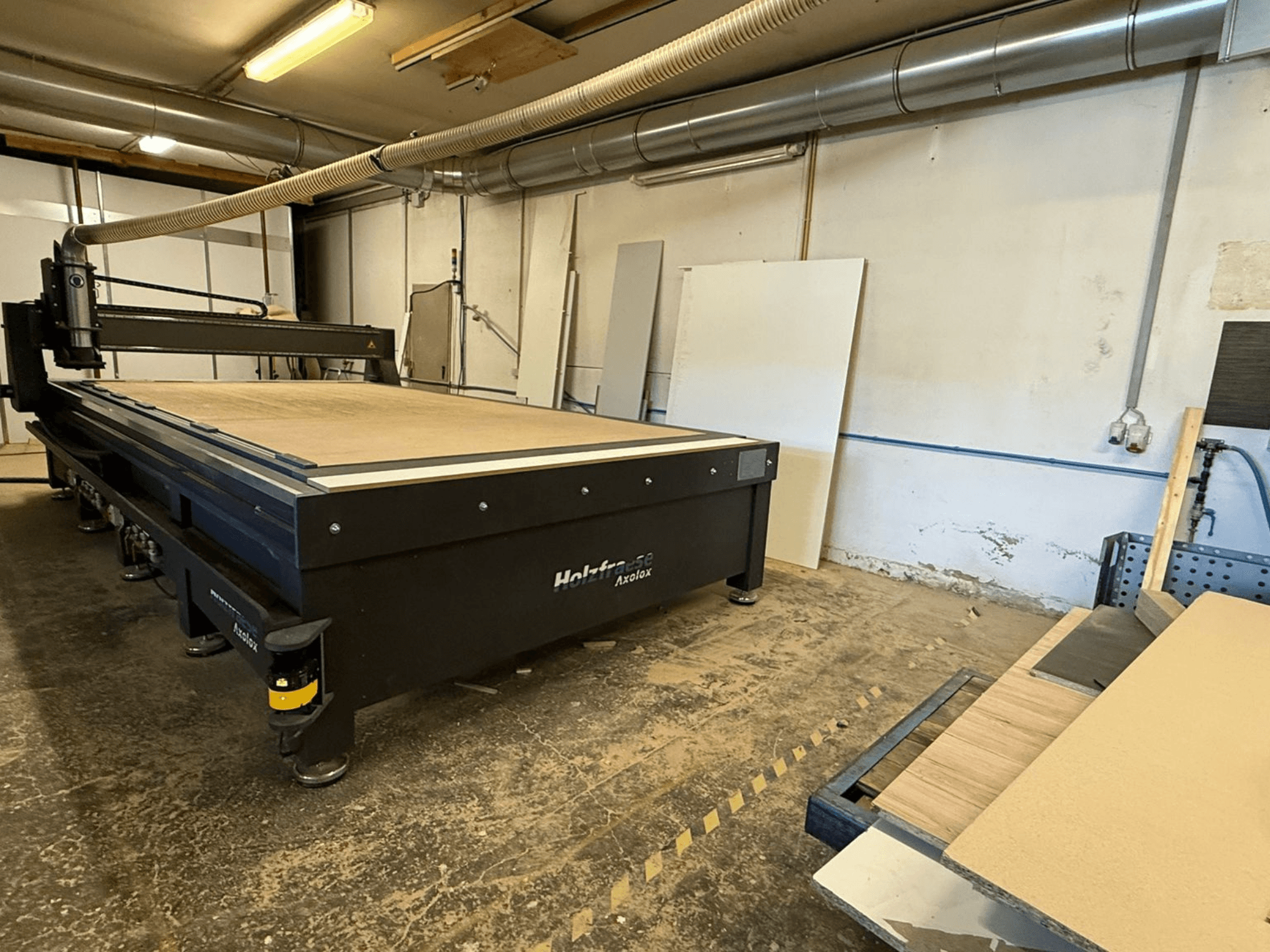 "Holz-Her Axos 5" CNC maršrutizatorius dirbtuvėse, vaizdas iš priekio, ant kurio matyti darbo stalas, dulkių ištraukimo sistema ir šalia esančios medinės plokštės.