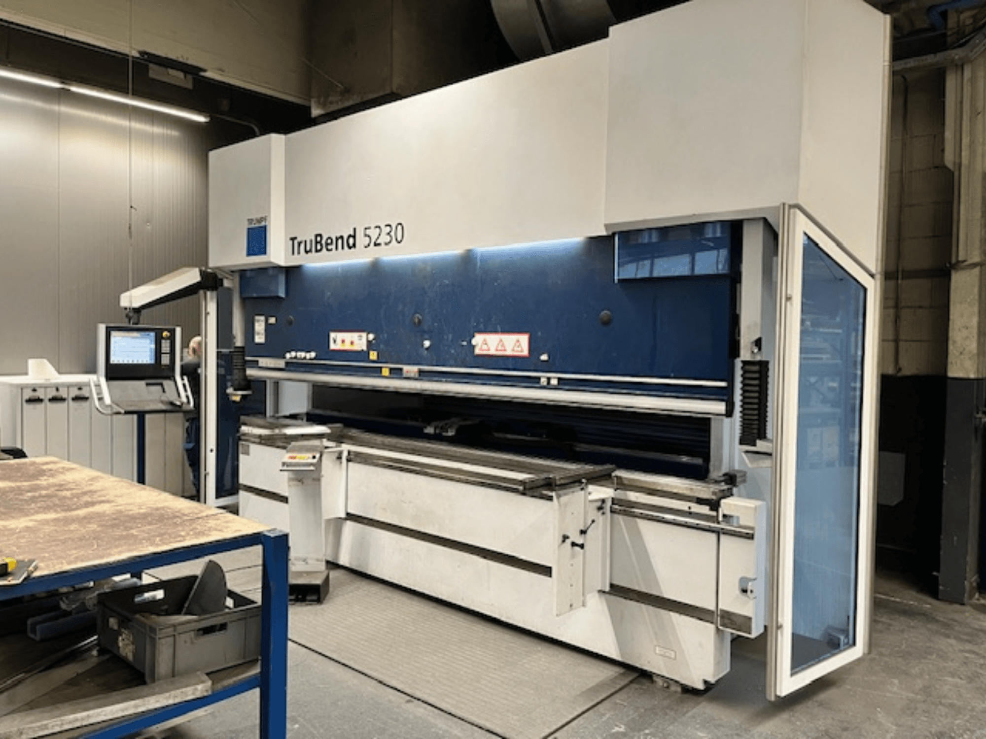 Lenkimo staklės "TruBend 5230" pramoninėje aplinkoje; priekinis vaizdas, kuriame matomas valdymo skydelis, saugos elementai ir darbo stalas.
