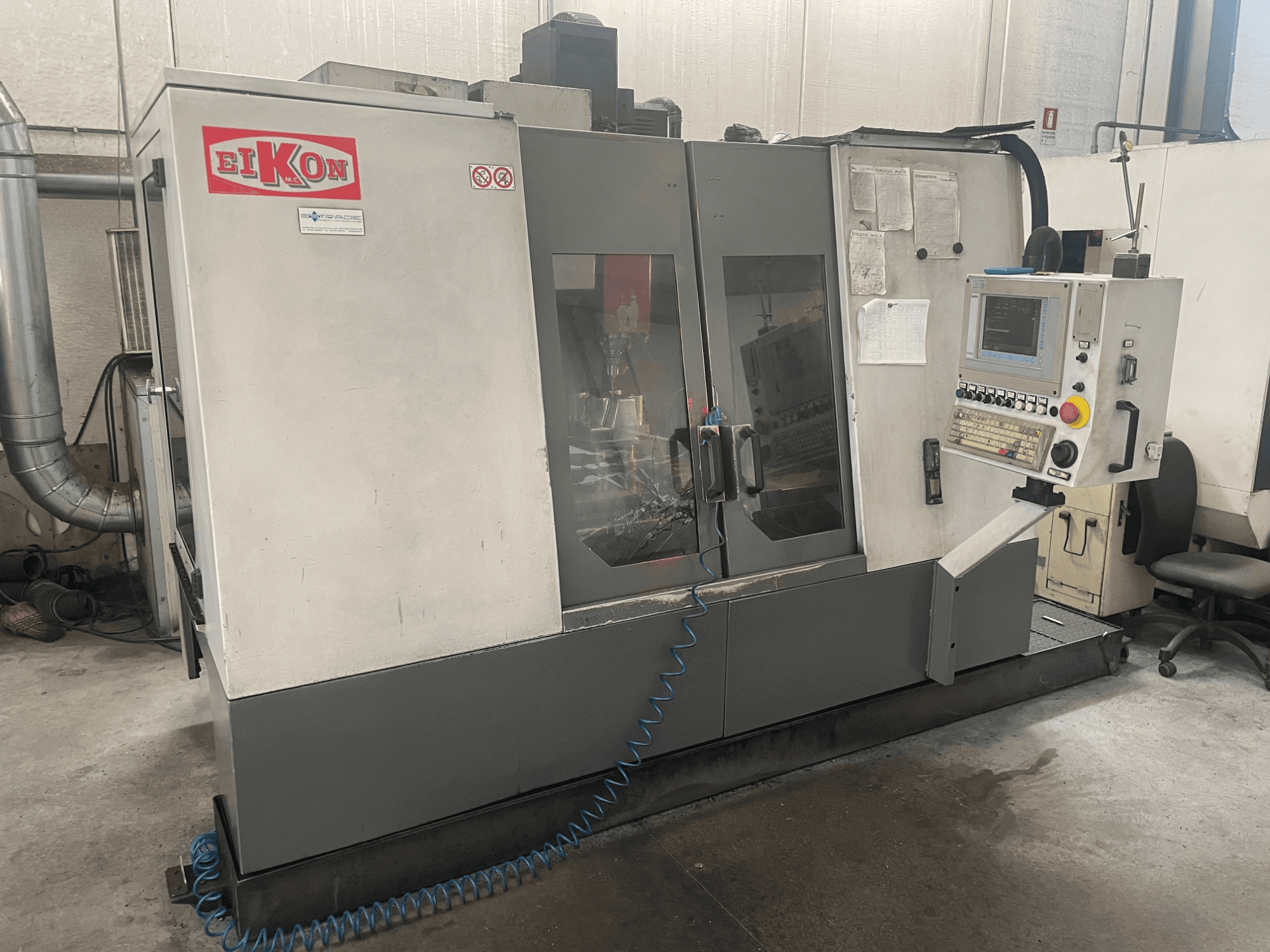 EIKON CNC staklės, žiūrint iš priekio, su valdymo pultu, didelėmis stiklinėmis durimis ir mėlyna oro tiekimo žarna ant grindų.